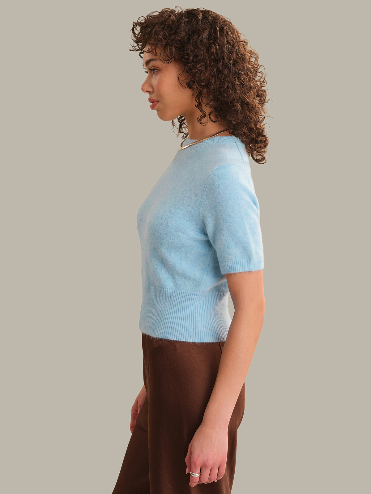 A woman with curly brown hair is shown in profile wearing a light blue short-sleeve knit top and brown pants against a solid beige background.