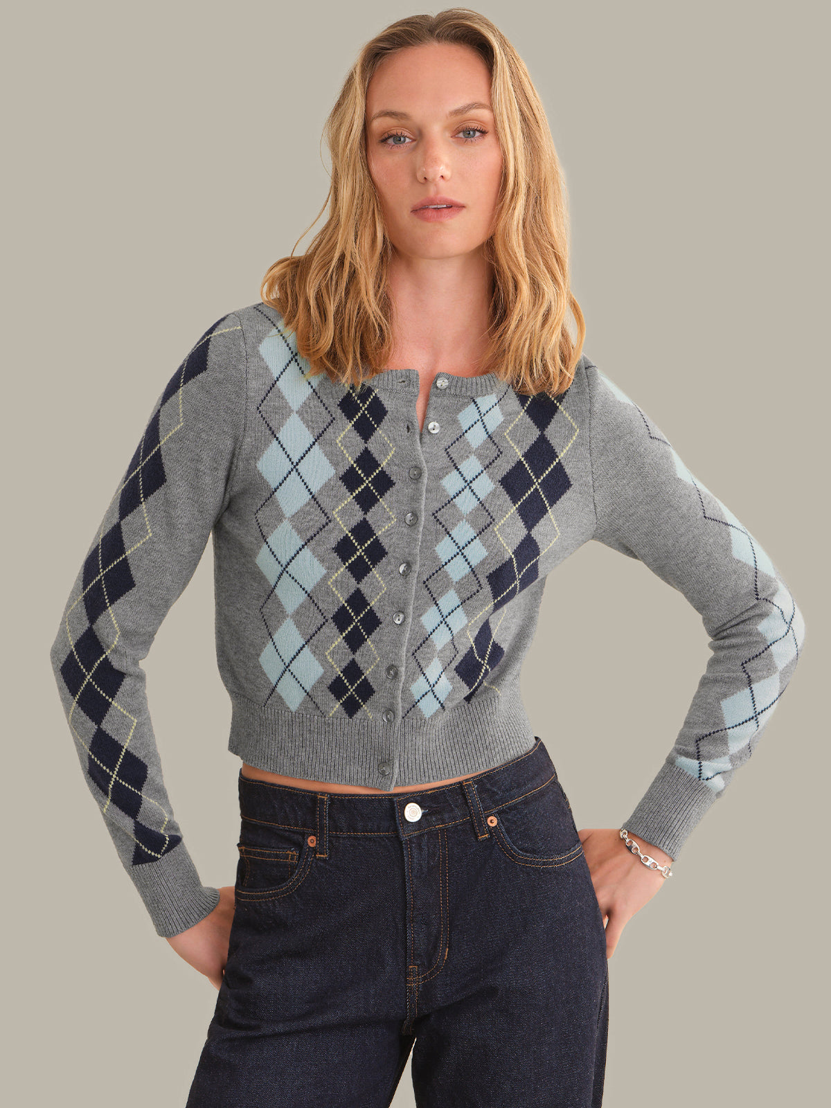 A woman modeling a gray cropped button-front cardigan with an argyle pattern in navy, light blue, and beige, paired with dark blue jeans against a solid taupe background.
