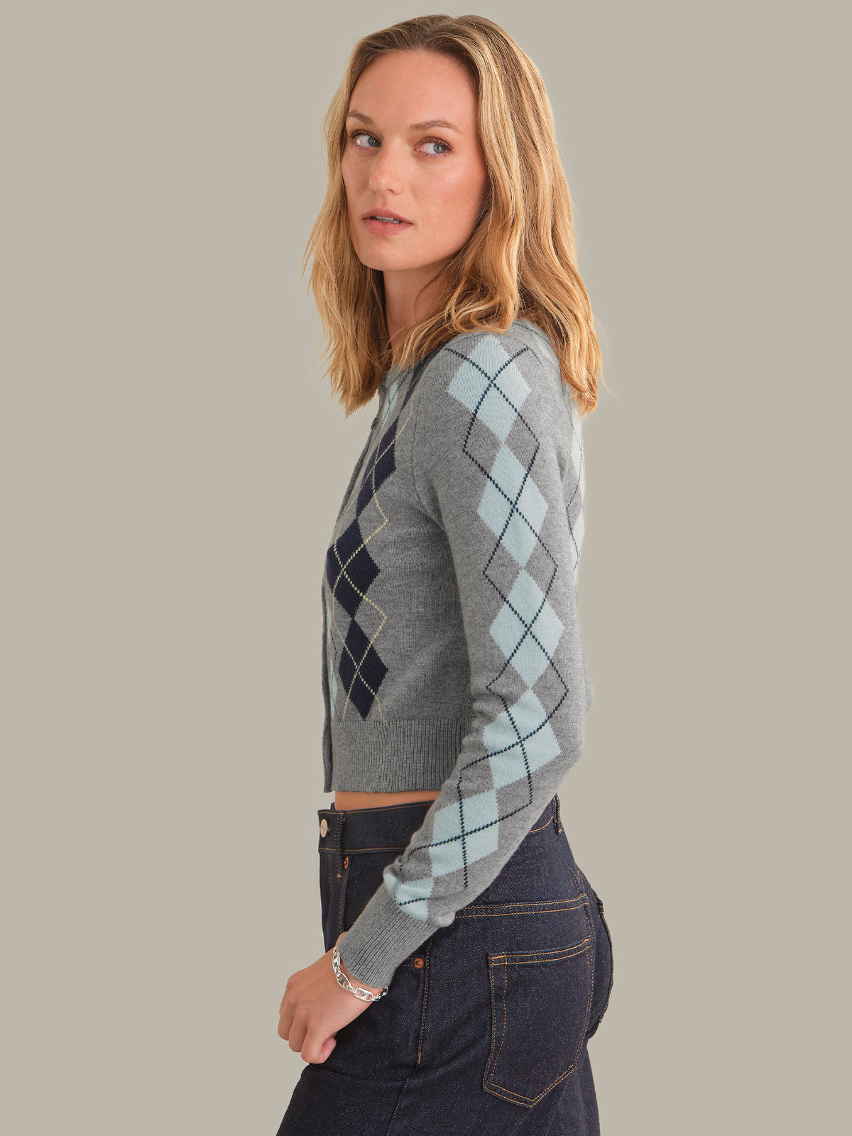A woman modeling a gray cropped long-sleeve sweater with an argyle pattern in navy and light blue, paired with dark blue jeans against a solid gray background.