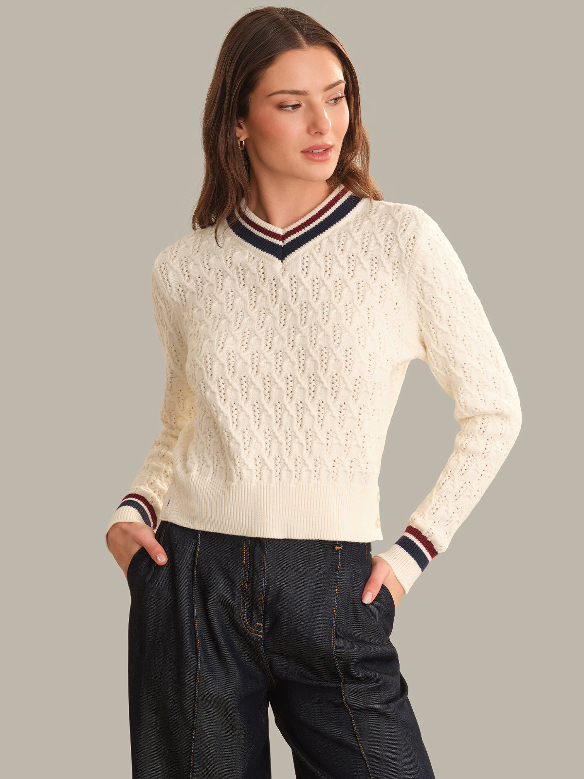A woman modeling a cream-colored cable-knit sweater with a navy and red striped V-neck collar and matching striped cuffs, paired with dark jeans against a solid gray background.