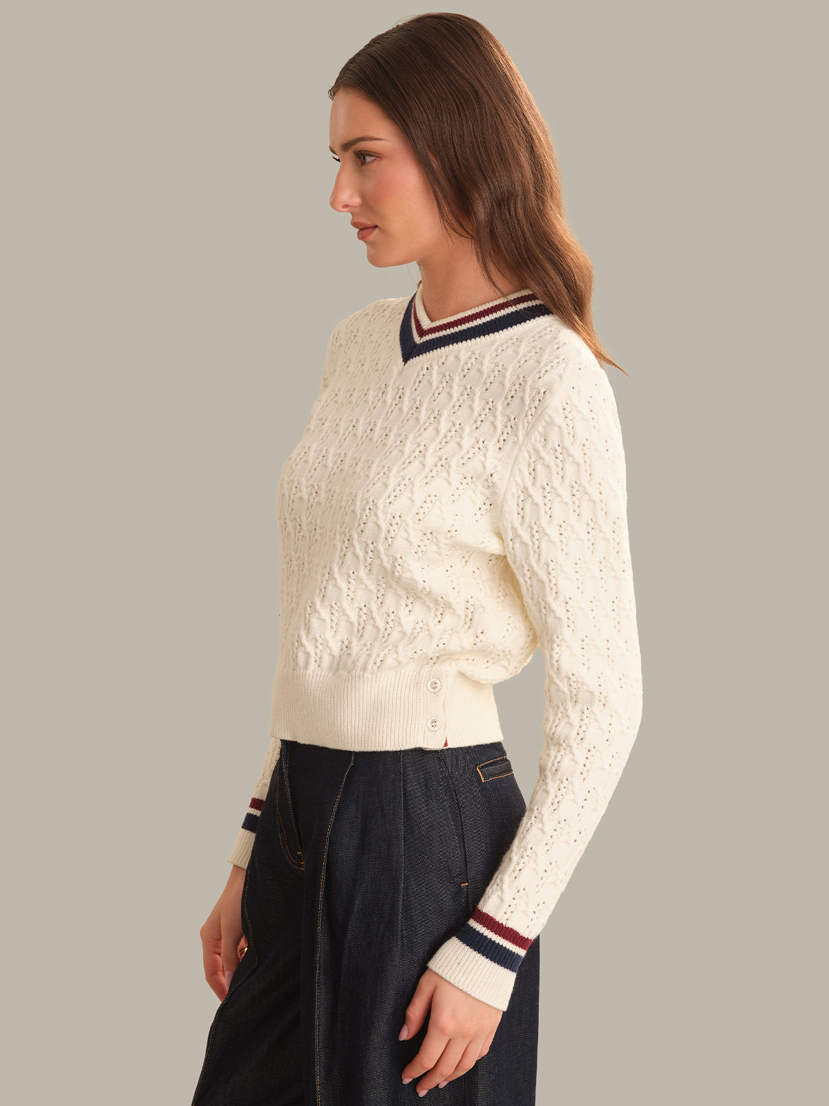 A woman shown in side profile wearing a cream cable-knit sweater with navy and red striped v-neck and cuff trim, paired with dark navy pants, against a solid light gray background.