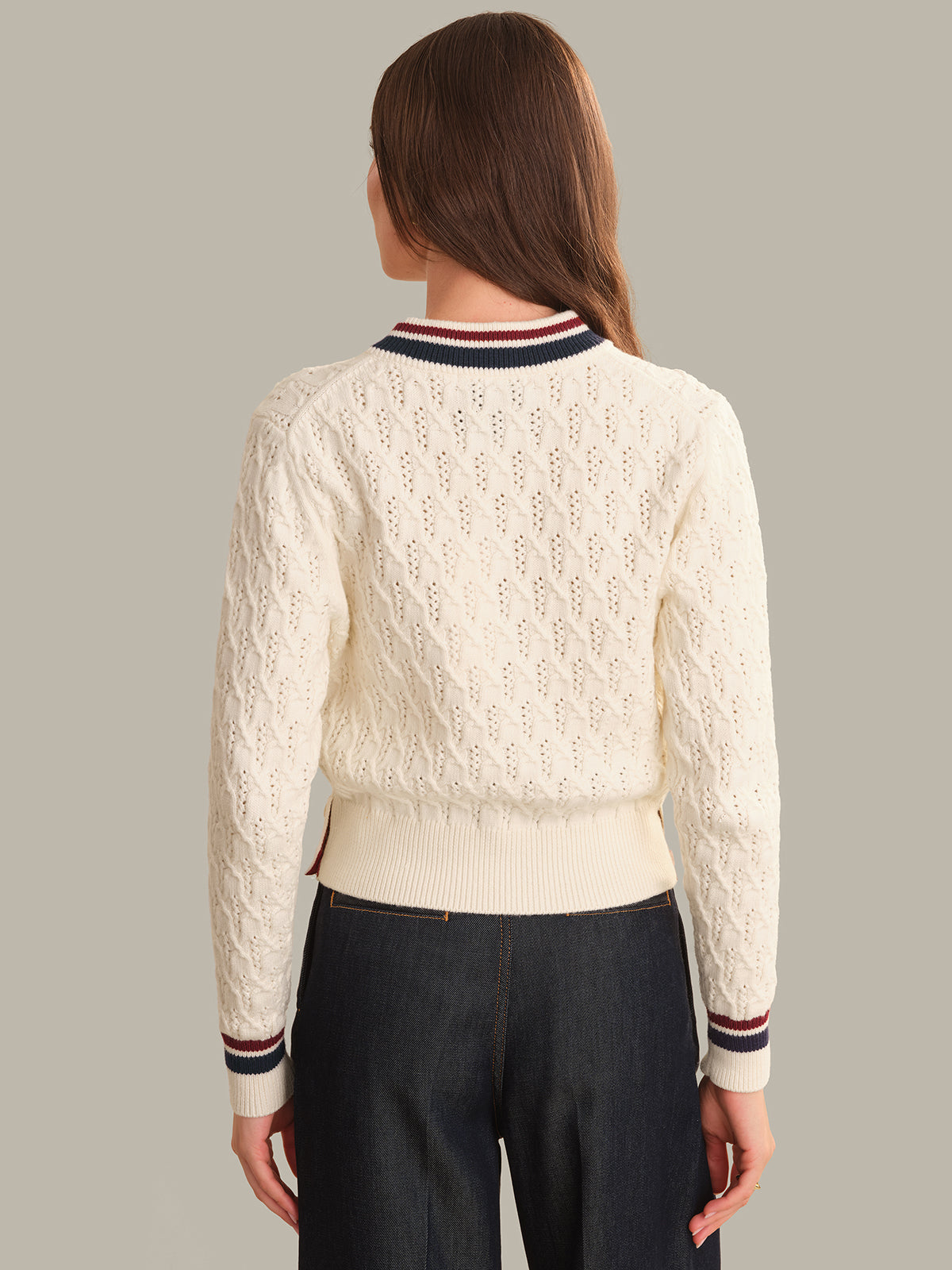 A woman viewed from the back wearing a cream cable-knit cropped sweater with navy, red, and white striped ribbed collar, hem, and cuffs, paired with dark jeans against a solid taupe background.