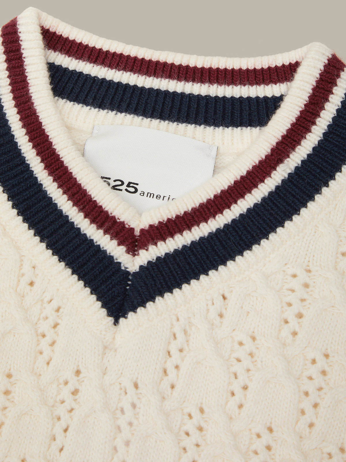 Close-up of a cream cable-knit sweater with a V-neck collar featuring navy blue and burgundy striped ribbing. A white neck label with '525 america' is visible.