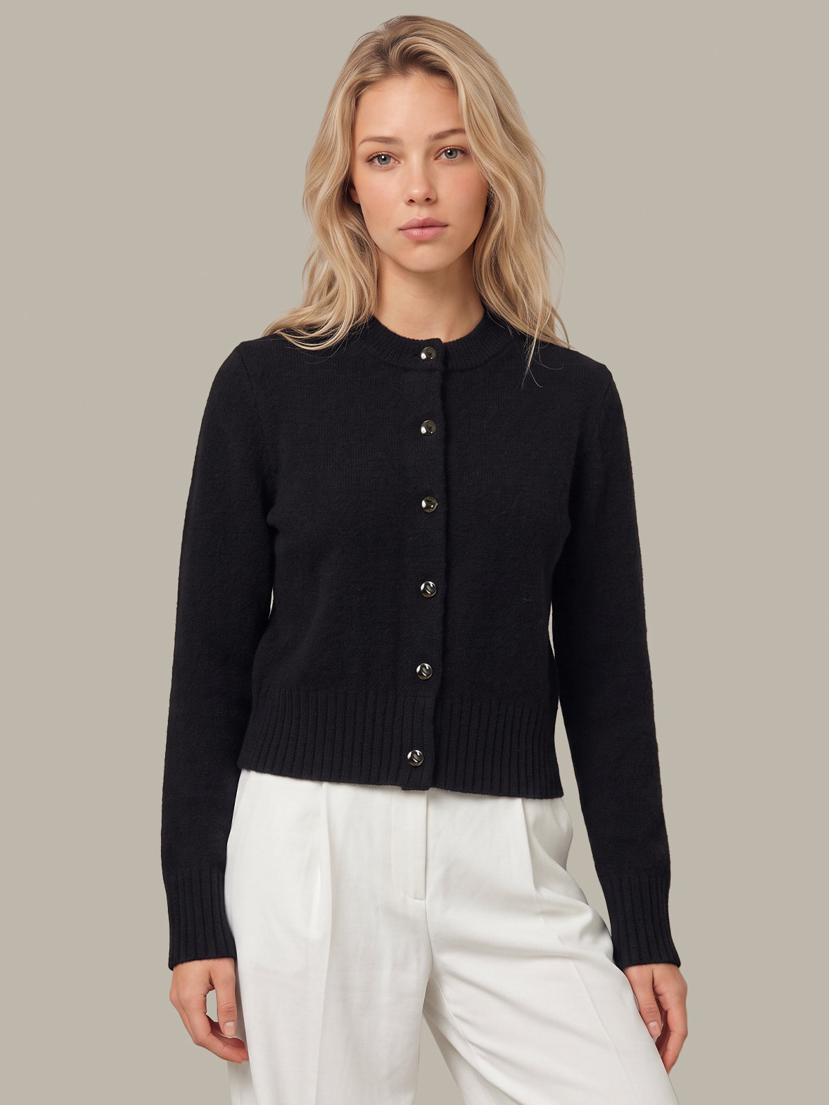 A blonde woman wearing a black long-sleeve button-front cardigan and white trousers, standing against a solid light gray studio background.