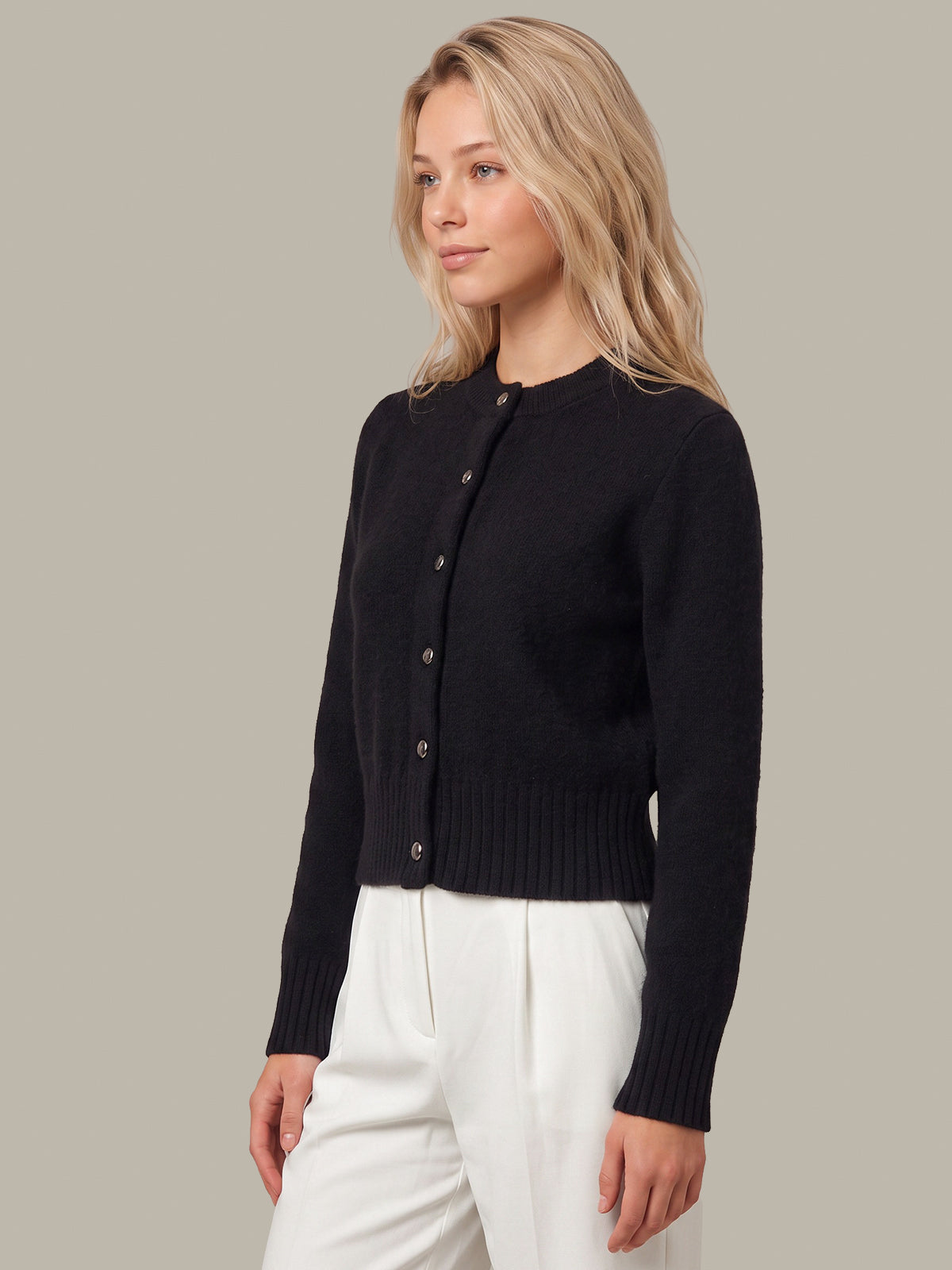 A woman wearing a black long-sleeve button-front cardigan and white trousers, photographed against a plain warm gray studio background.
