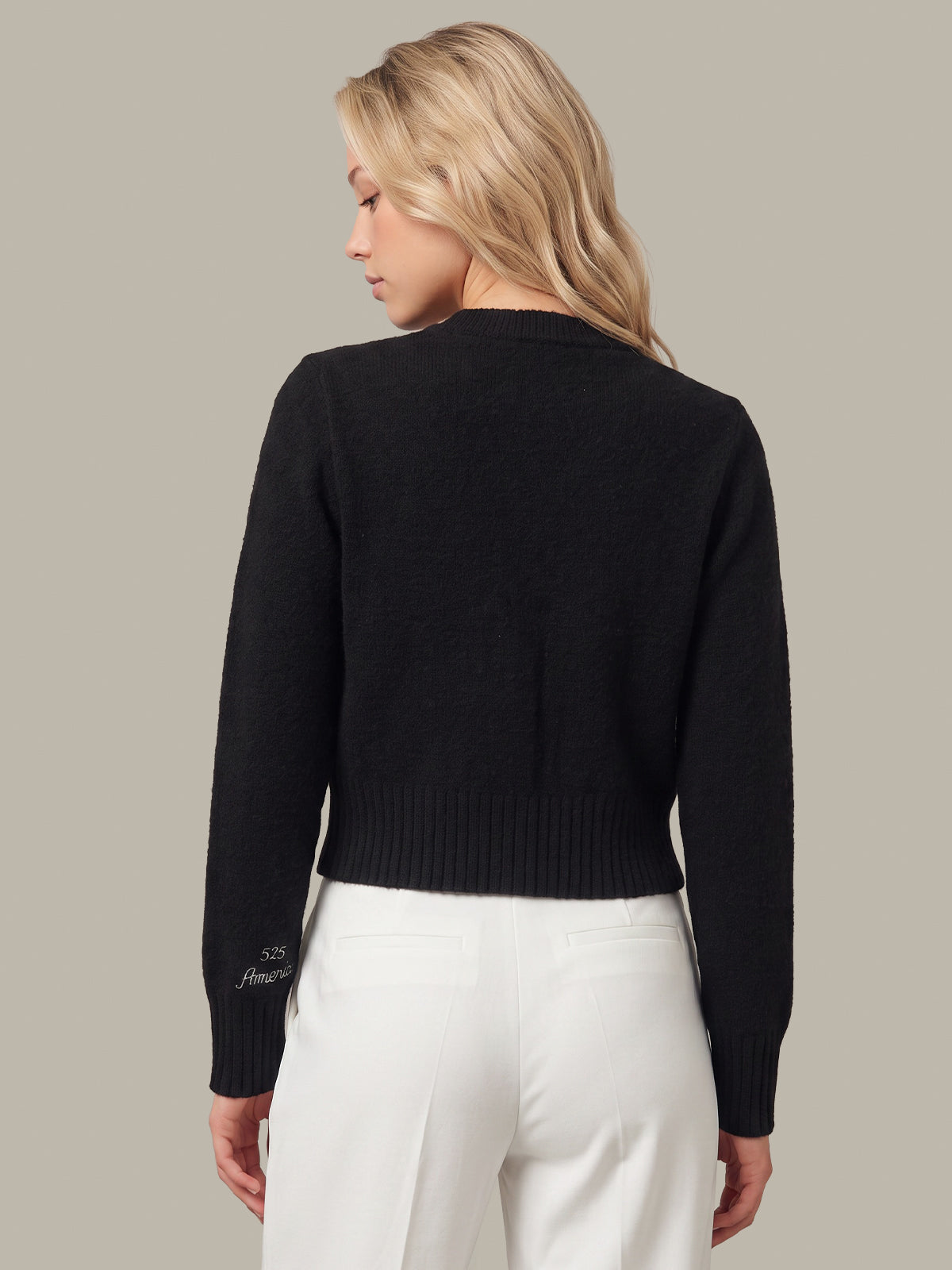 A woman shown from the back wearing a black cropped knit sweater and white trousers, photographed against a plain warm gray studio background.