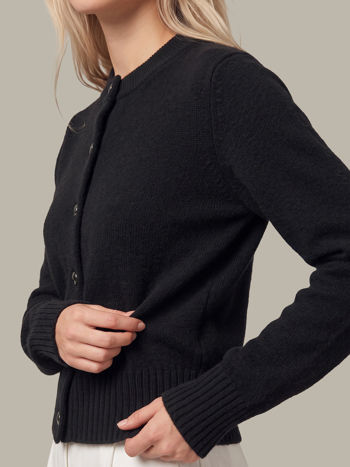 A close-up of a woman wearing a black ribbed knit cardigan sweater with a button front, shown against a plain warm gray background.