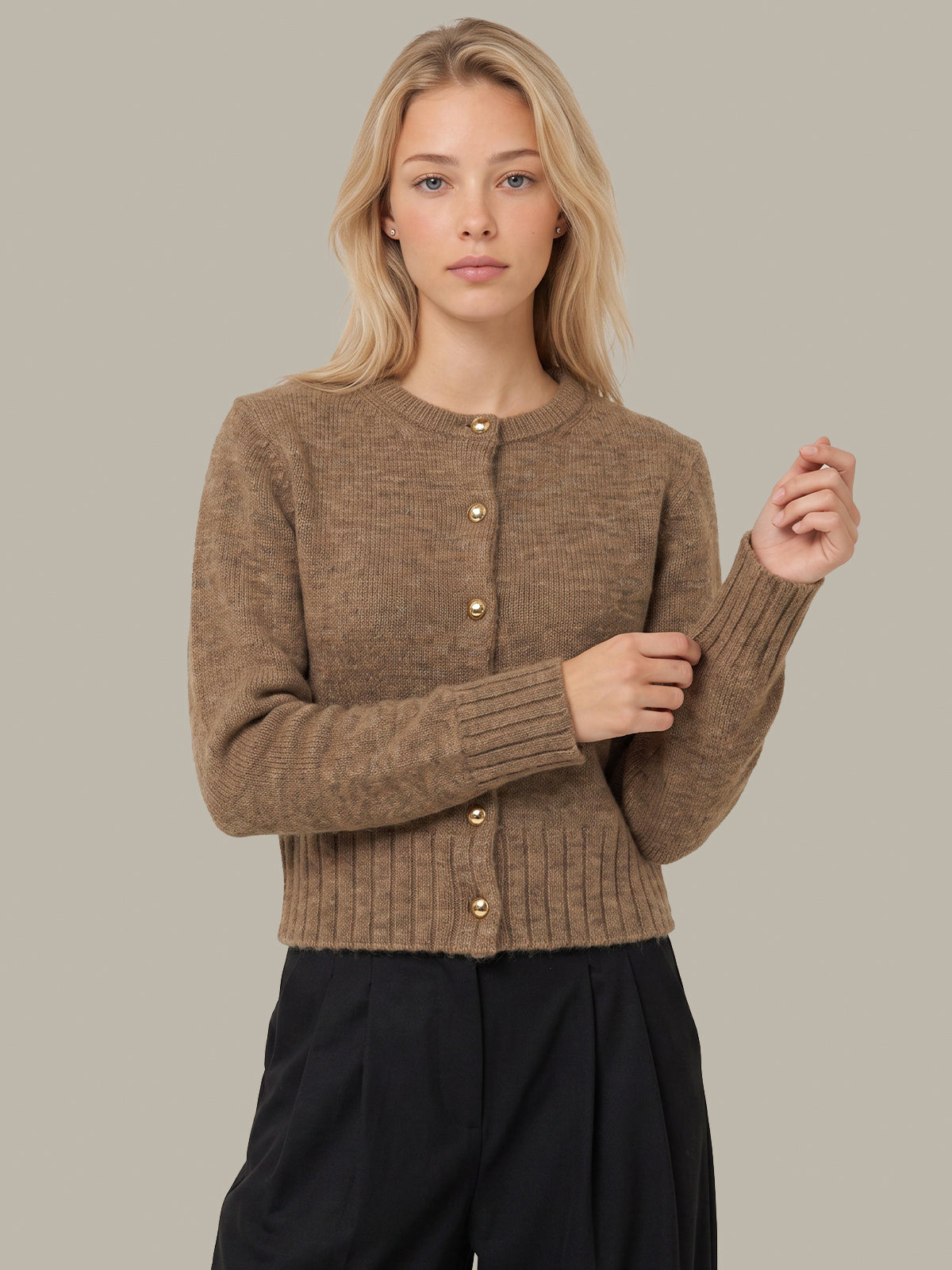 A woman modeling a cropped, long-sleeve ribbed knit cardigan in a warm taupe-brown color with gold-tone buttons, paired with black trousers, against a solid light gray background.