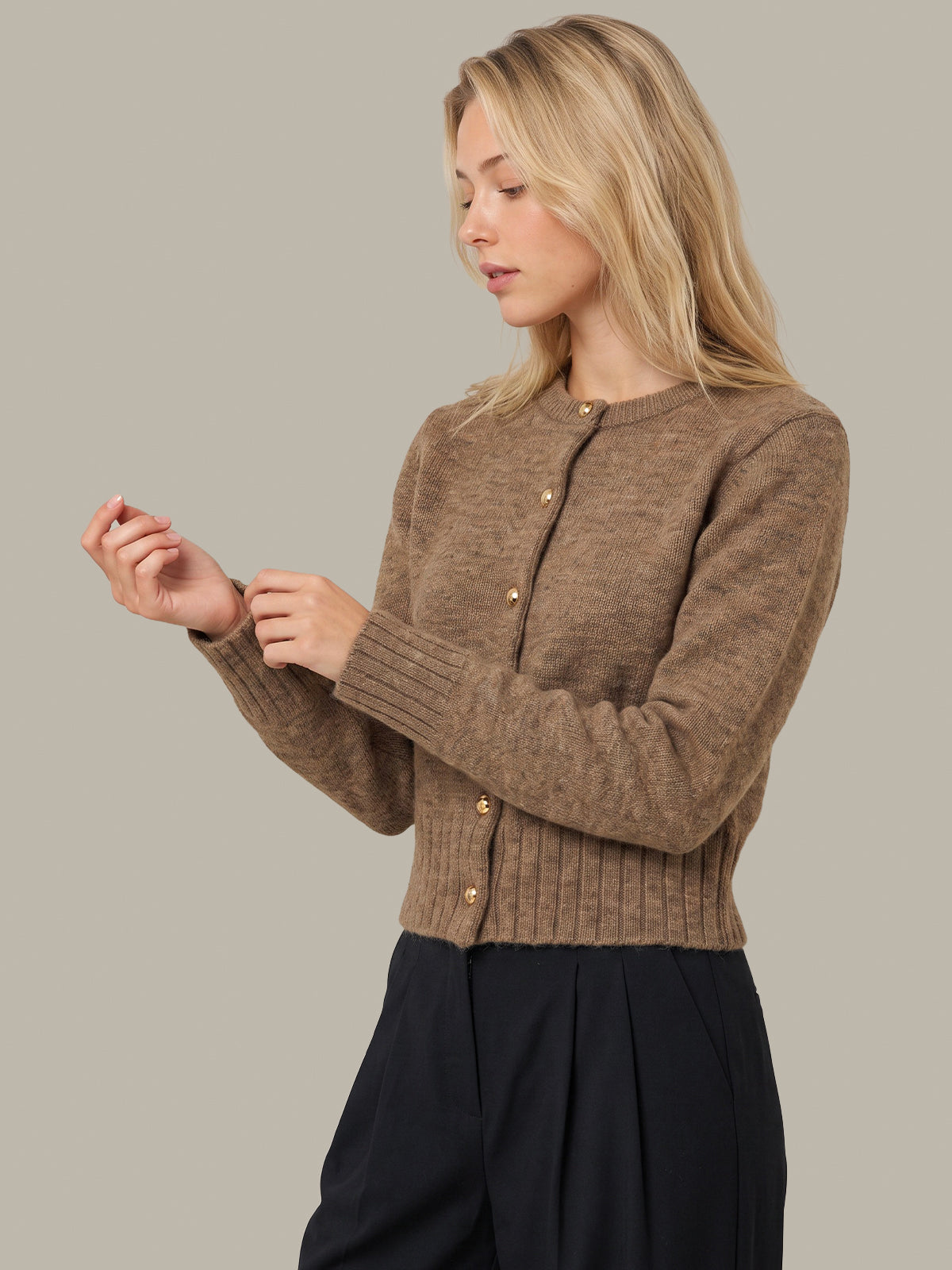 A woman wearing a cropped, ribbed-knit brown cardigan with gold-tone buttons and dark navy trousers, shown against a solid light gray studio background.