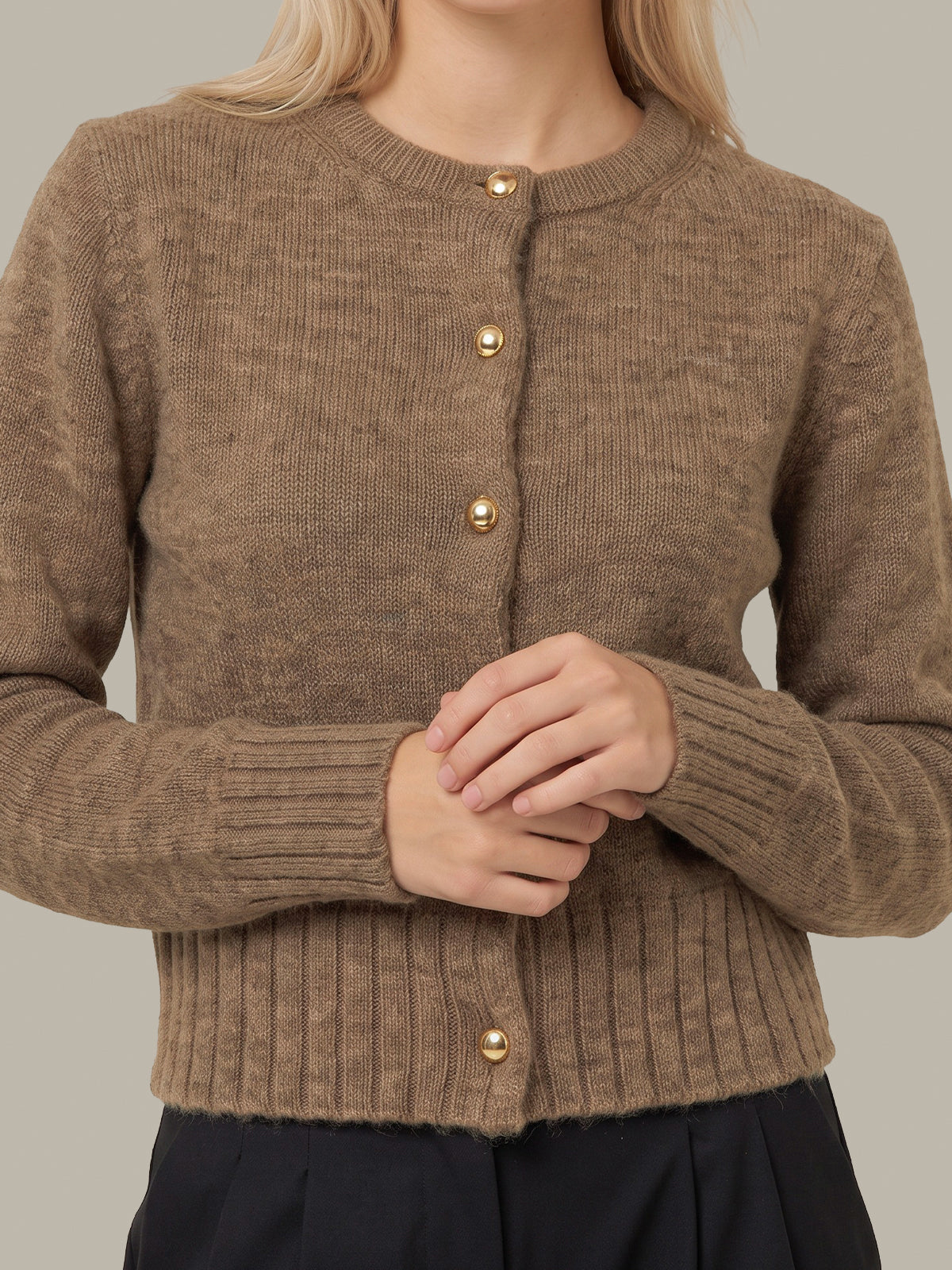 A close-up of a woman wearing a brown ribbed knit cardigan sweater with gold buttons, paired with dark navy bottoms, against a solid light gray background.