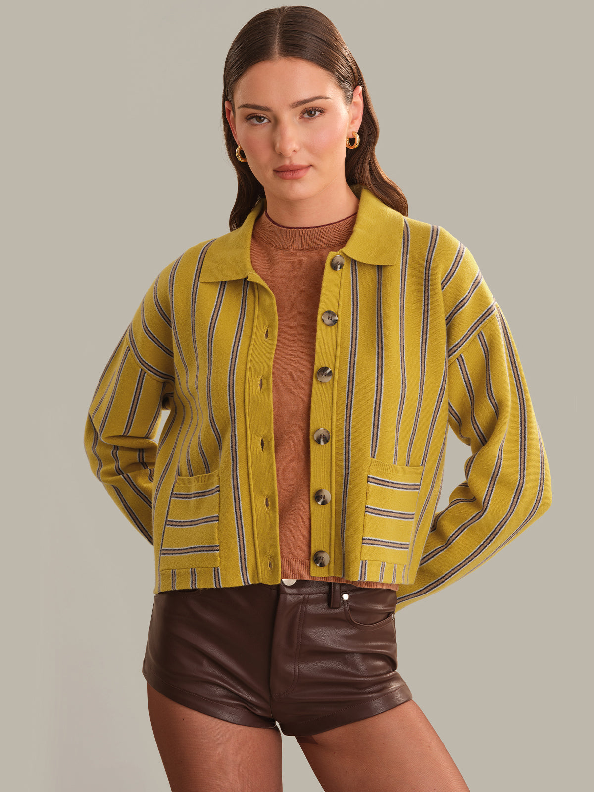 A woman modeling a cropped yellow button-front cardigan with thin dark vertical stripes, worn over a brown mock-neck top and dark brown faux-leather shorts, against a solid light gray studio background.