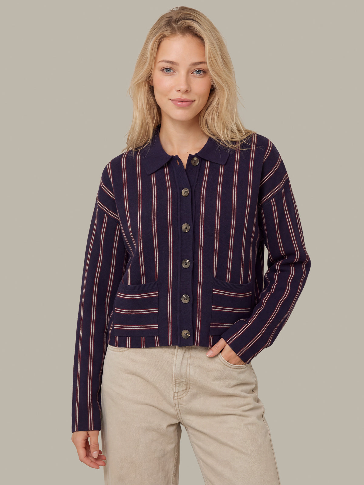 A woman modeling a navy blue button-up cardigan with thin pink vertical stripes, paired with beige pants, against a solid taupe background.