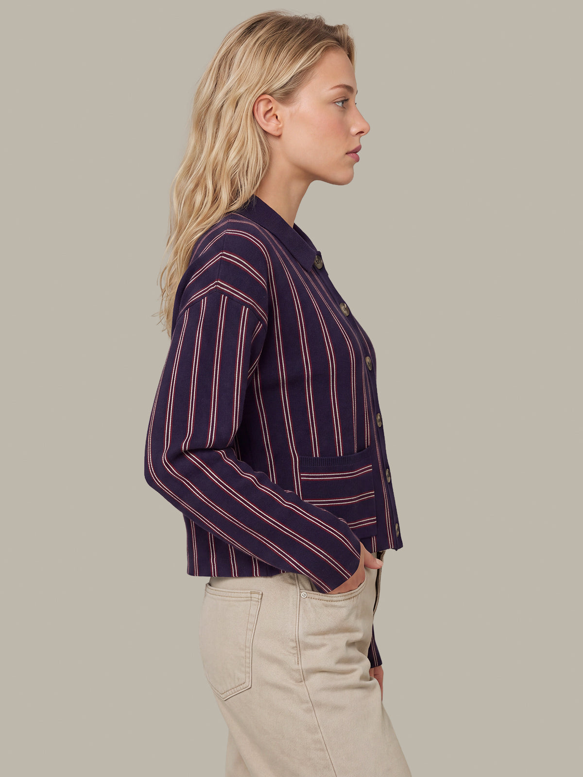 A woman shown in side profile wearing a cropped long-sleeve button-front blazer with vertical navy, burgundy, and beige stripes, paired with light beige pants, against a solid warm gray studio background.