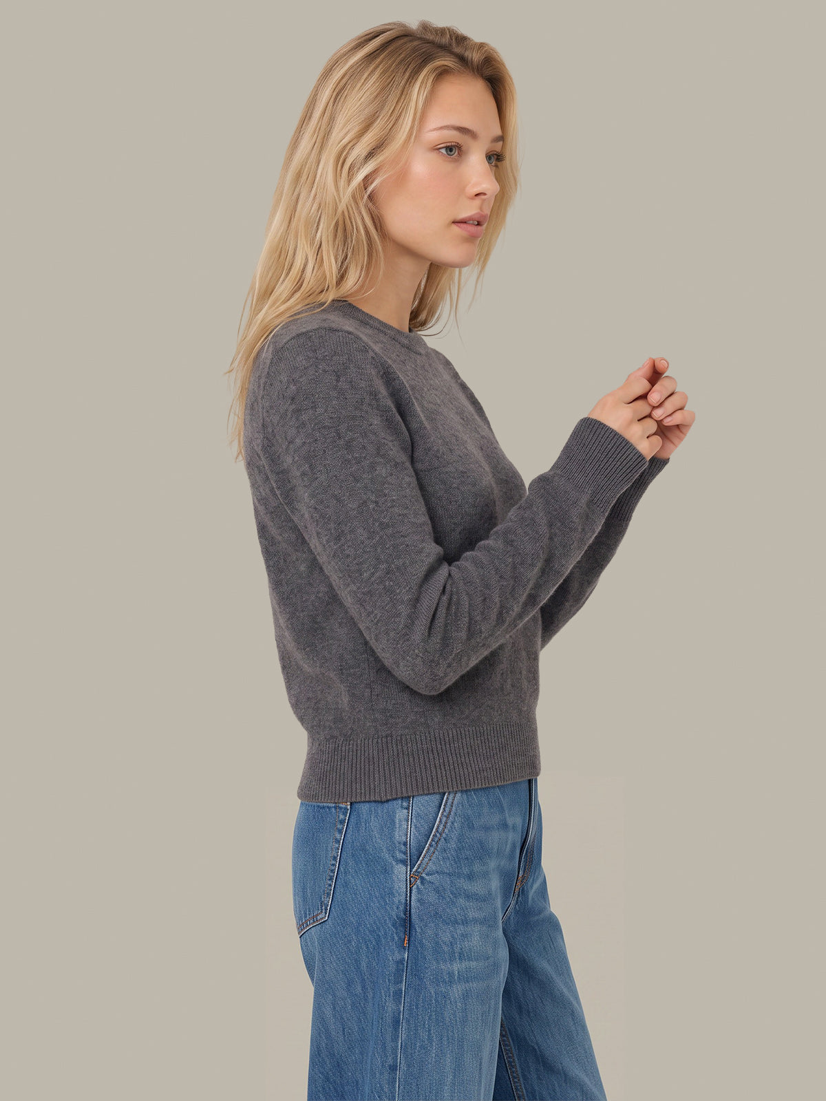 A woman wearing a dark gray knit crewneck sweater and blue jeans, shown from a side profile against a solid light gray studio background.