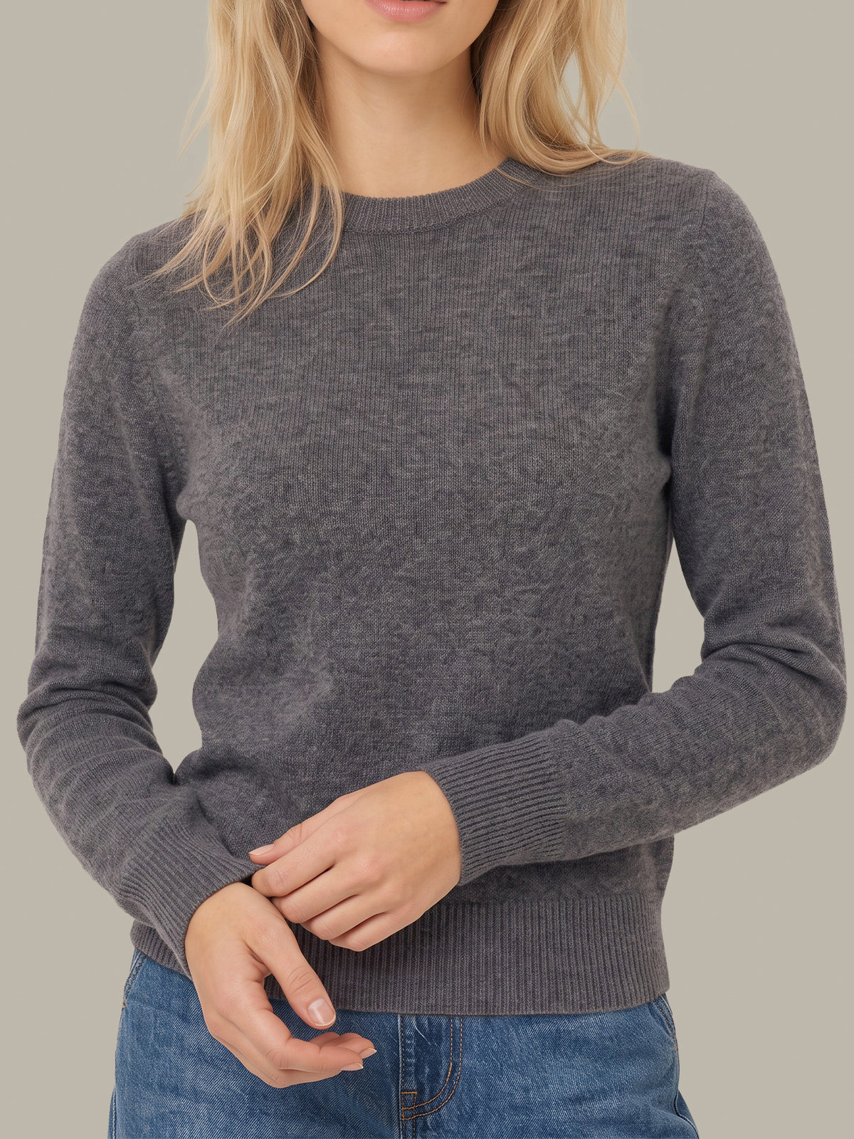 A woman wearing a dark gray heathered knit crewneck sweater with ribbed cuffs and hem, paired with blue jeans, shown against a solid light gray background.