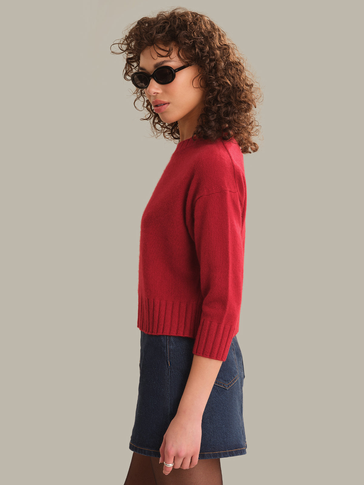 A woman with curly brown hair wearing a red knit sweater and a blue denim skirt, shown in profile against a solid light gray background.
