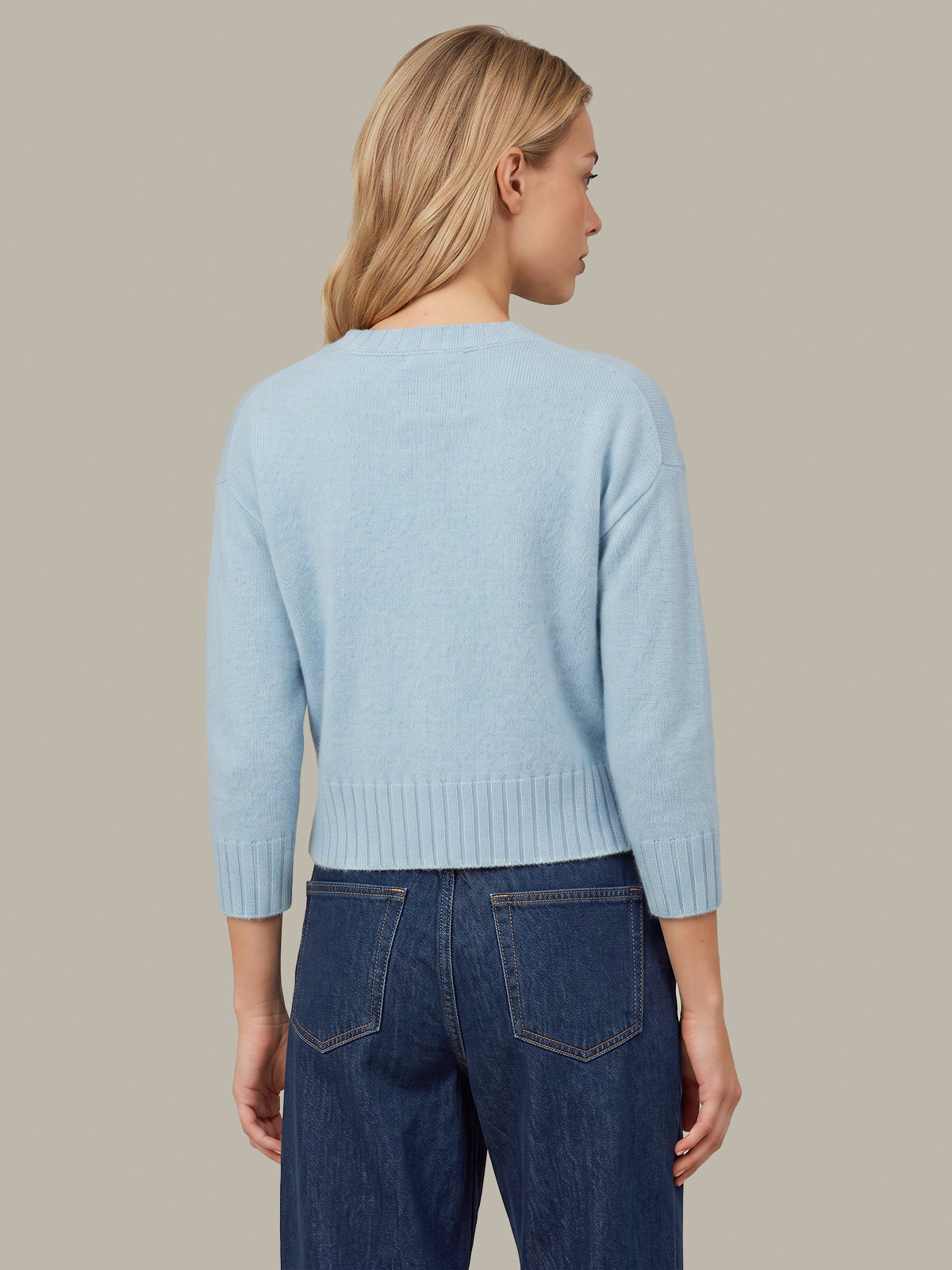 A woman shown from the back wearing a light blue knit sweater and dark blue jeans against a solid taupe background.
