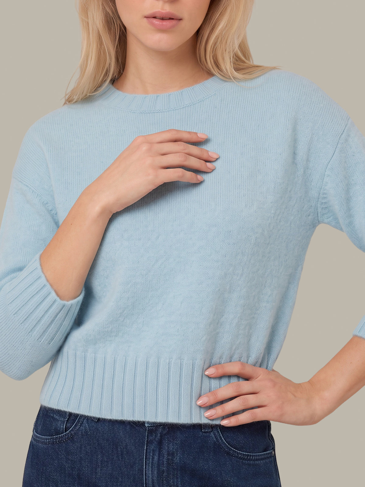 A woman wearing a light blue ribbed knit sweater with three-quarter sleeves and a crew neck, paired with dark blue jeans, against a solid beige background.
