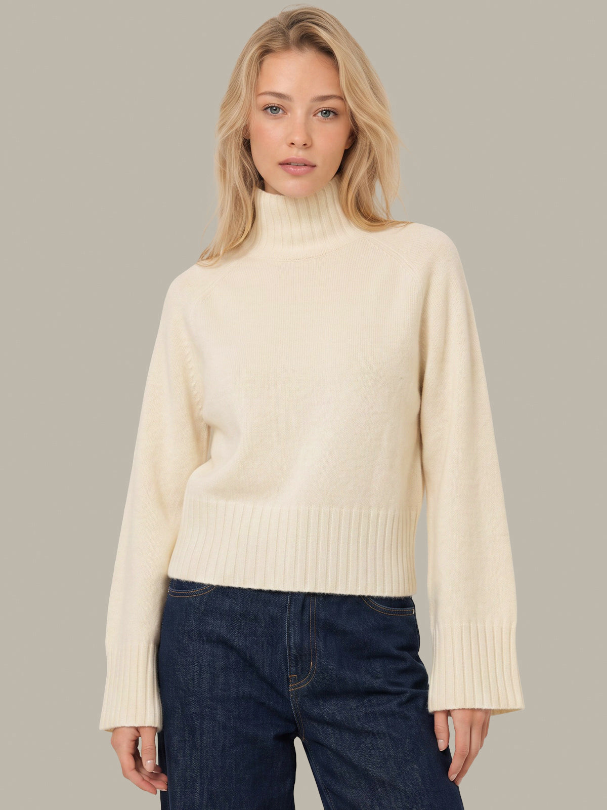 A blonde woman wearing a cream-colored ribbed turtleneck sweater and dark blue jeans, standing against a solid gray studio background.