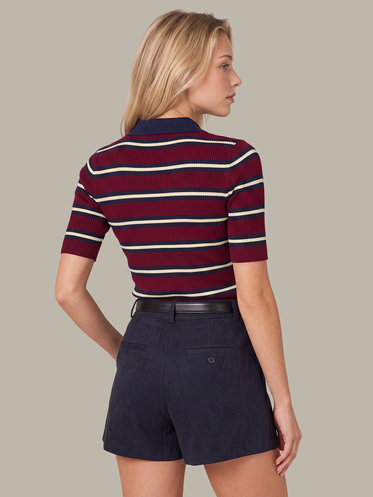 A woman shown from the back wearing a short-sleeve ribbed knit polo shirt with horizontal navy, burgundy, and cream stripes, paired with dark navy shorts, against a solid taupe background.