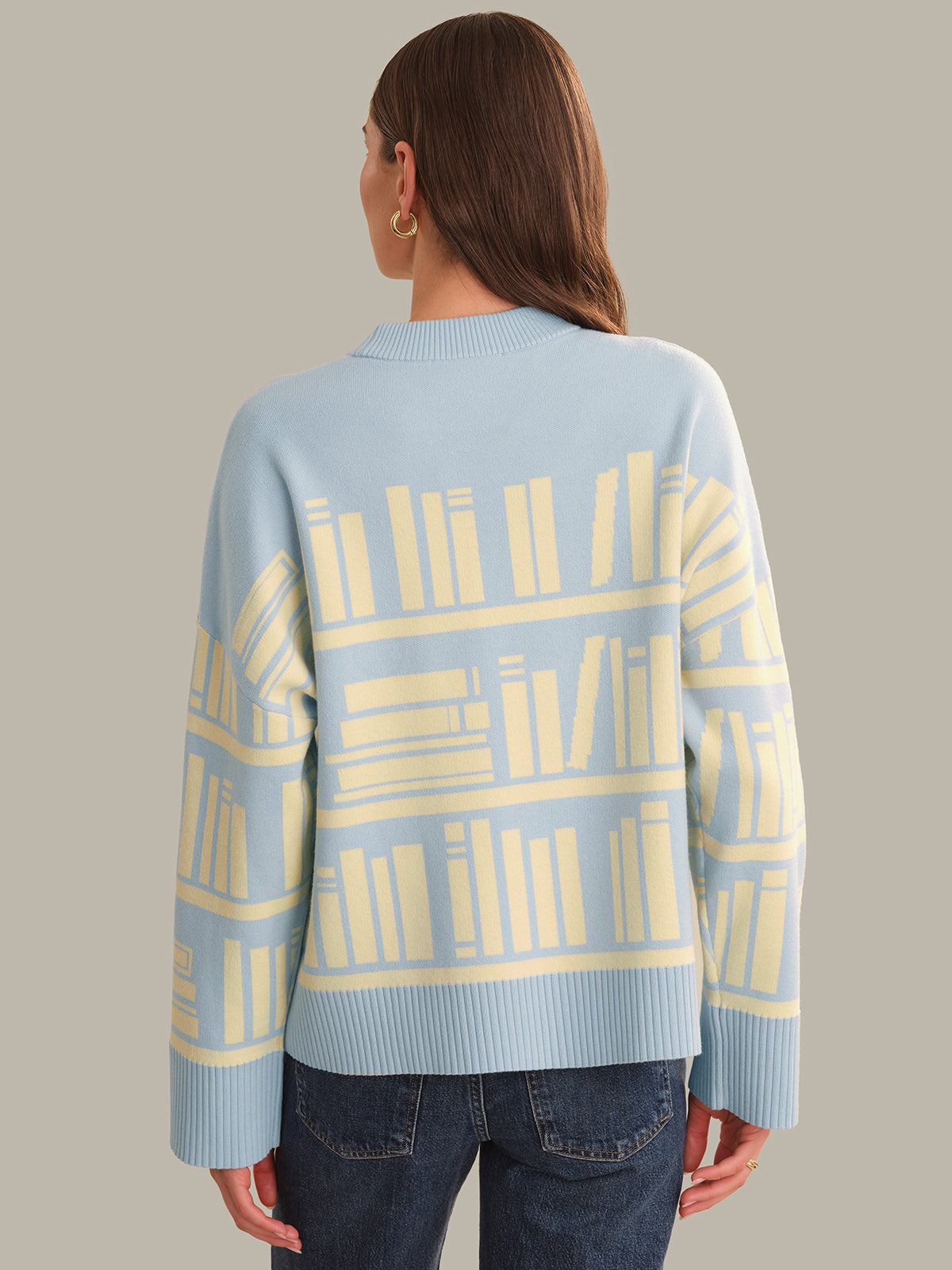 A woman viewed from the back wearing a light blue knit sweater with a pale yellow bookshelf pattern, paired with dark blue jeans, against a solid beige background.
