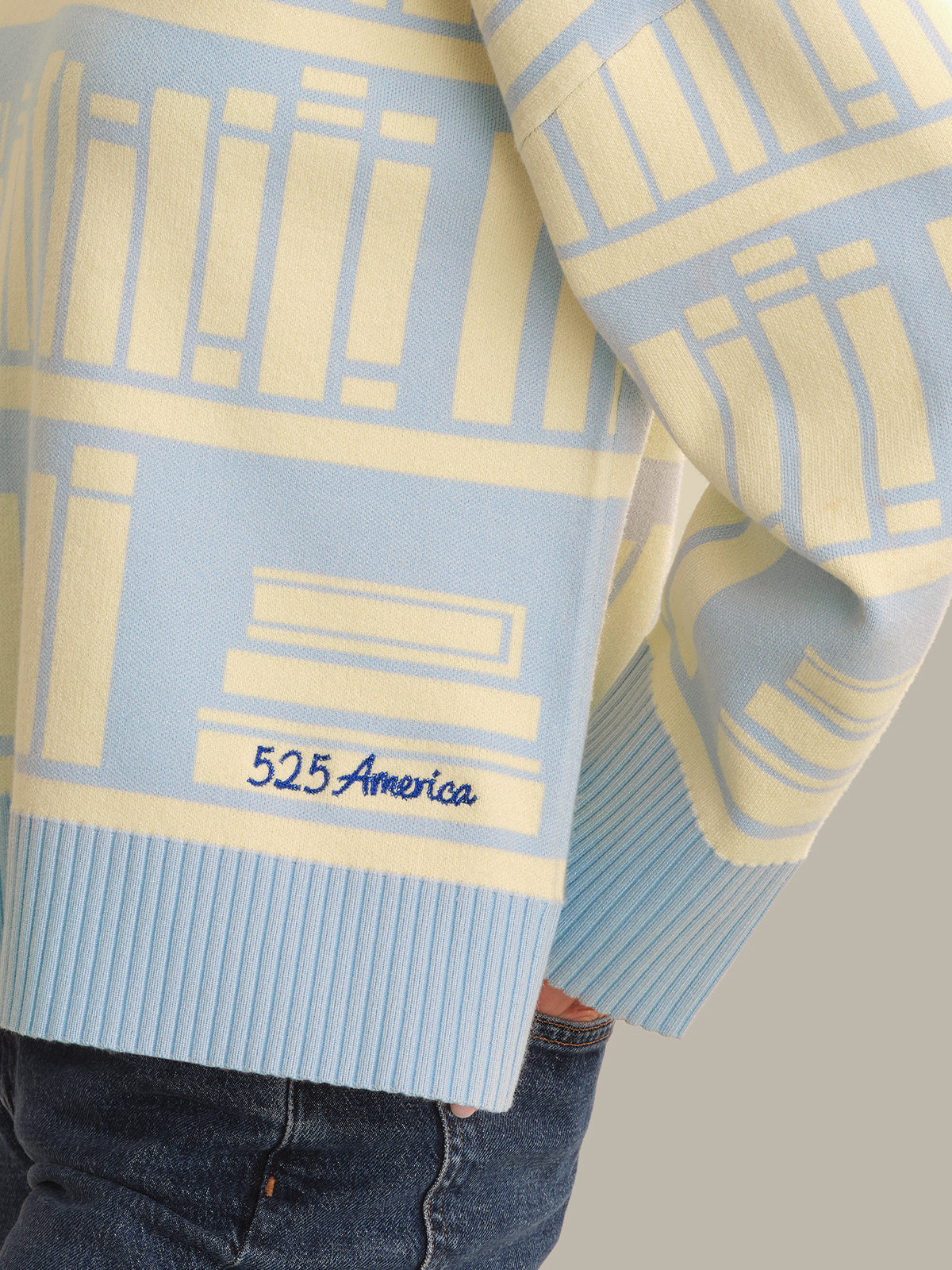 A close-up of a light blue and cream knit sweater featuring a geometric bookshelf pattern, ribbed cuffs and hem, and the brand name '525 America' embroidered in navy on the lower left front.