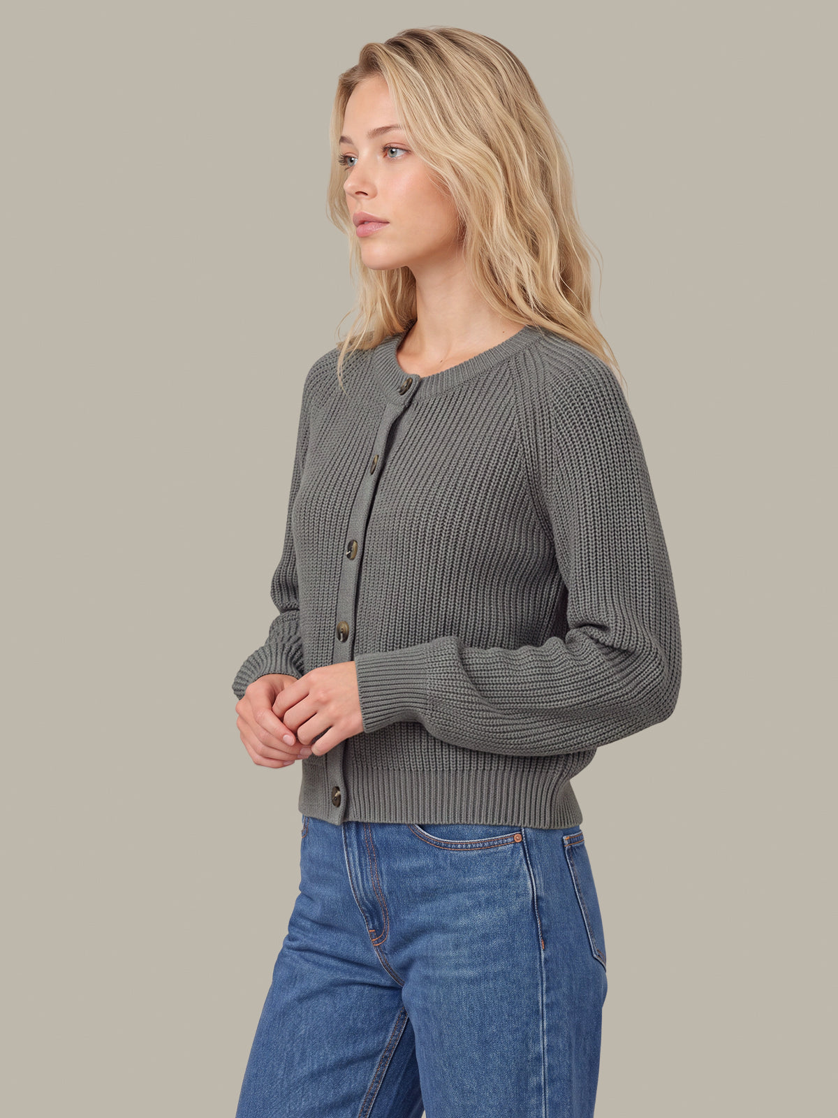 A woman wearing a dark gray ribbed knit cardigan sweater with brown buttons, paired with blue jeans, shown from a side profile against a solid beige background.