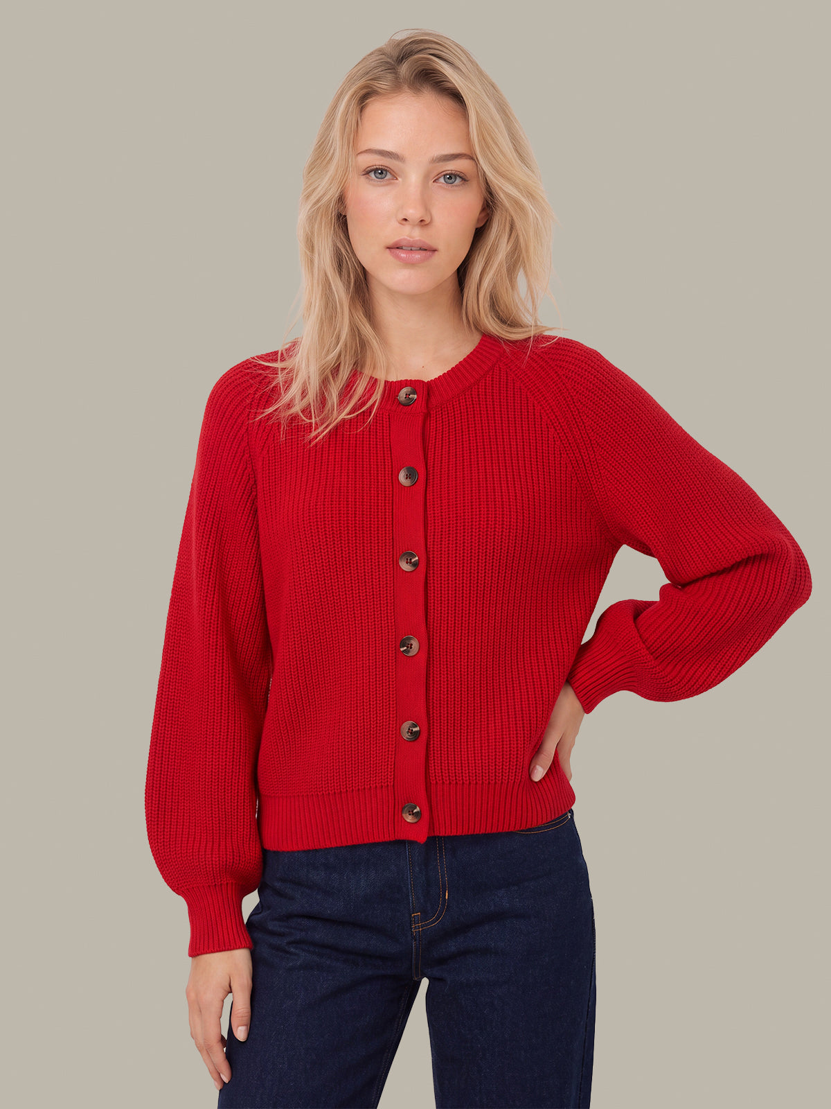 A woman modeling a bright red ribbed knit cardigan sweater with dark buttons, paired with dark blue jeans, against a solid light gray background.