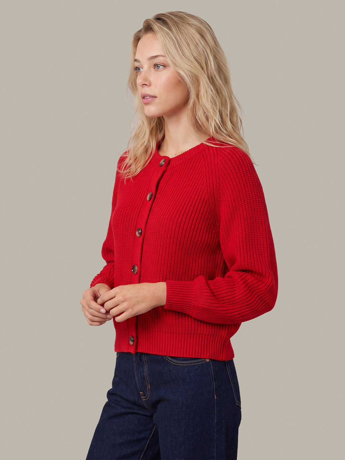 A woman wearing a bright red ribbed knit cardigan sweater with dark buttons, paired with dark blue jeans, photographed against a solid light gray background.