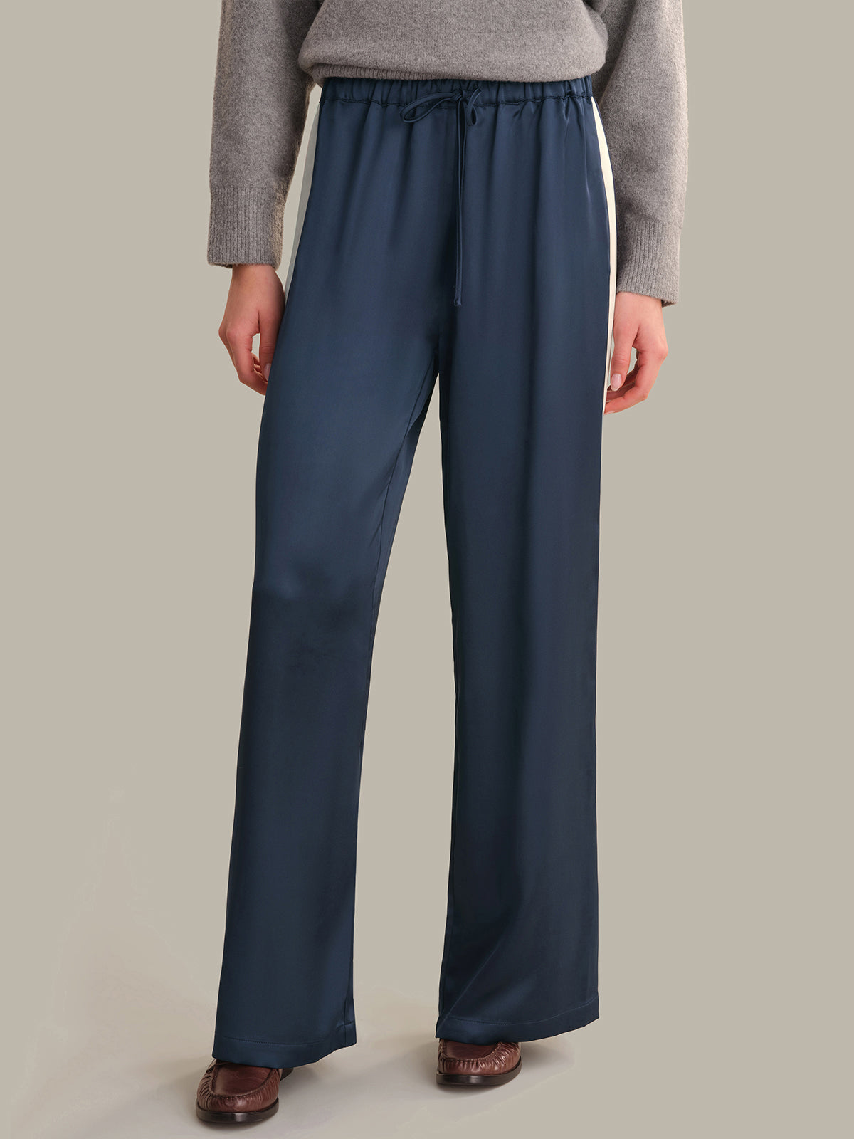 A person wearing dark navy wide-leg pants with white side stripes, a gray sweater, and brown shoes, standing against a plain beige studio background.