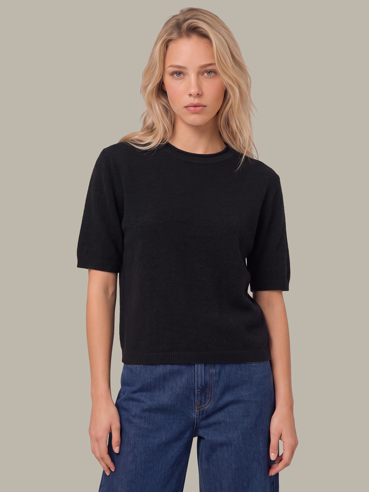 A woman modeling a black short-sleeve crewneck knit top with blue jeans, set against a solid neutral gray background.
