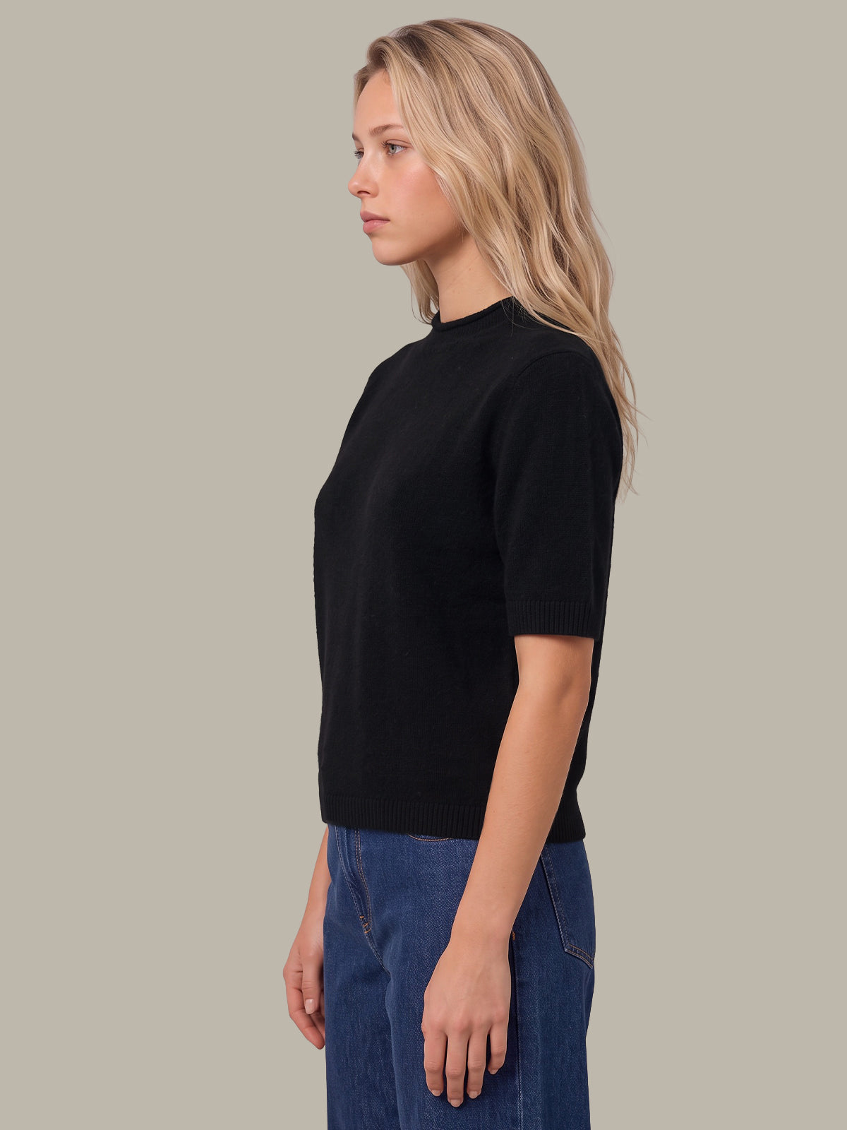 A woman in profile wearing a black short-sleeve knit top and blue jeans, photographed against a plain warm gray studio background.