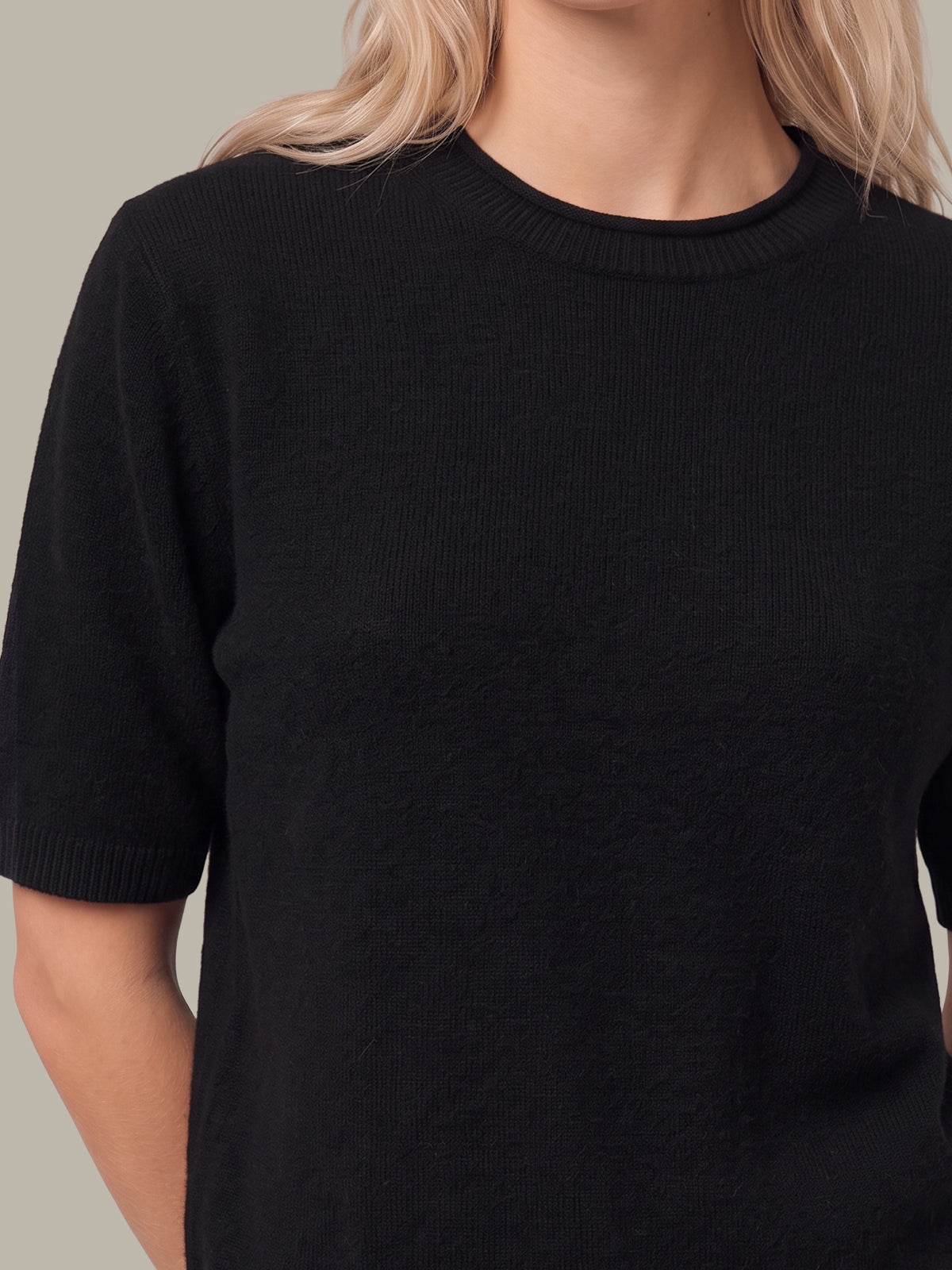 A close-up of a person wearing a black short-sleeve knit crewneck sweater against a plain light gray background.