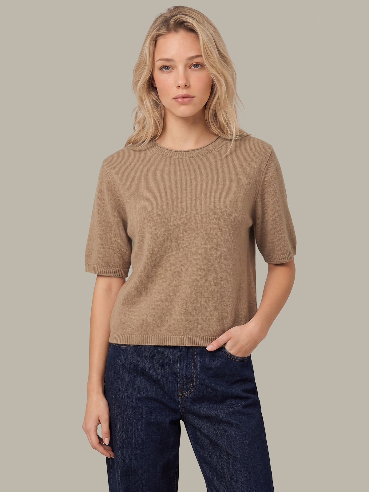 A woman modeling a short-sleeve crewneck knit sweater in a warm taupe-brown color, paired with dark blue jeans, against a solid light gray background.