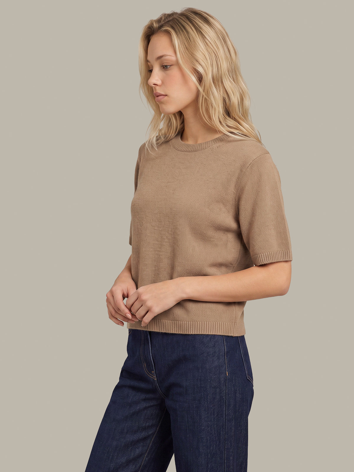A woman wearing a short-sleeve crewneck knit sweater in taupe and dark blue jeans, photographed against a solid light gray studio background.