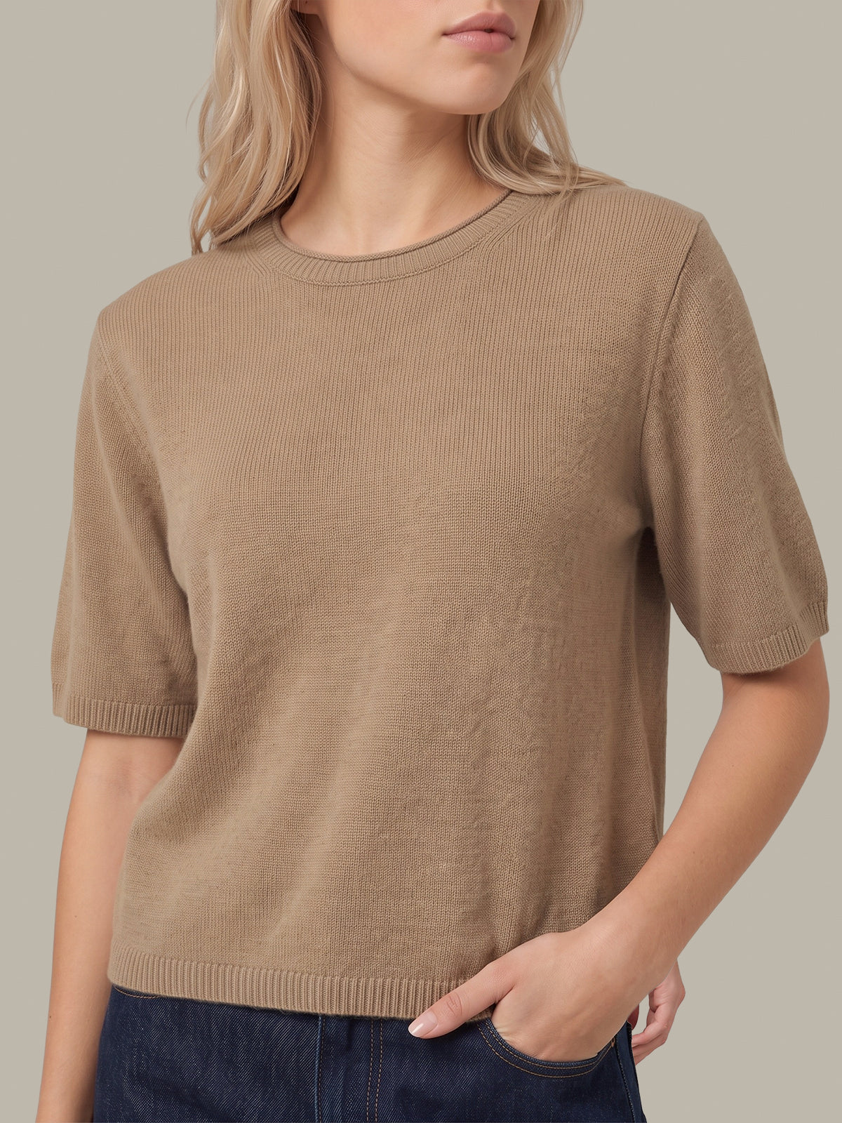 A woman wearing a short-sleeve ribbed knit sweater in a warm taupe-brown color, paired with dark blue jeans, against a solid neutral background.