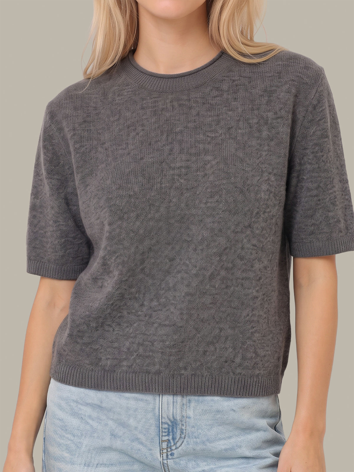 A woman wearing a dark gray short-sleeve knit sweater and light blue jeans, shown from the neck down against a plain beige background.