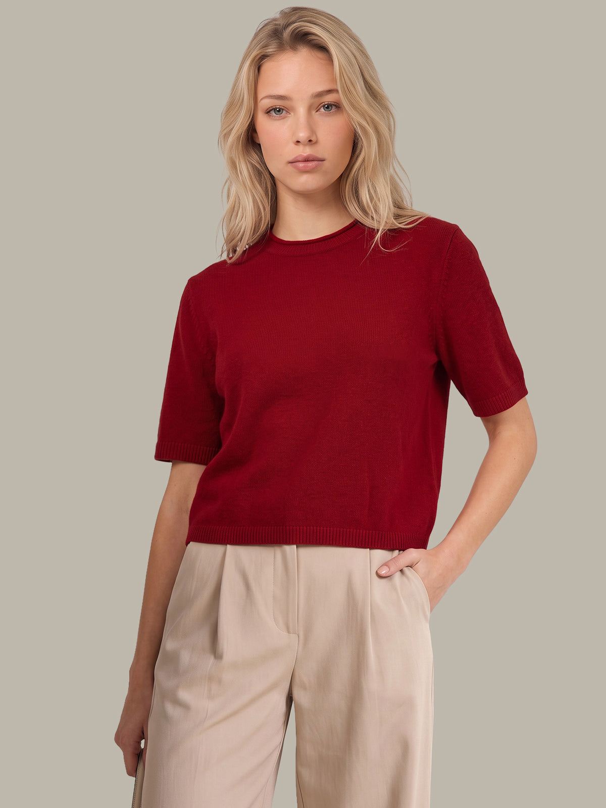 A blonde woman wearing a short-sleeve crewneck knit sweater in deep burgundy, paired with beige wide-leg trousers, against a solid light gray background.