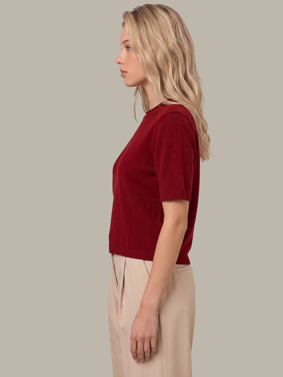 A woman in profile wearing a short-sleeve crewneck knit top in deep burgundy and beige trousers, set against a solid light gray background.