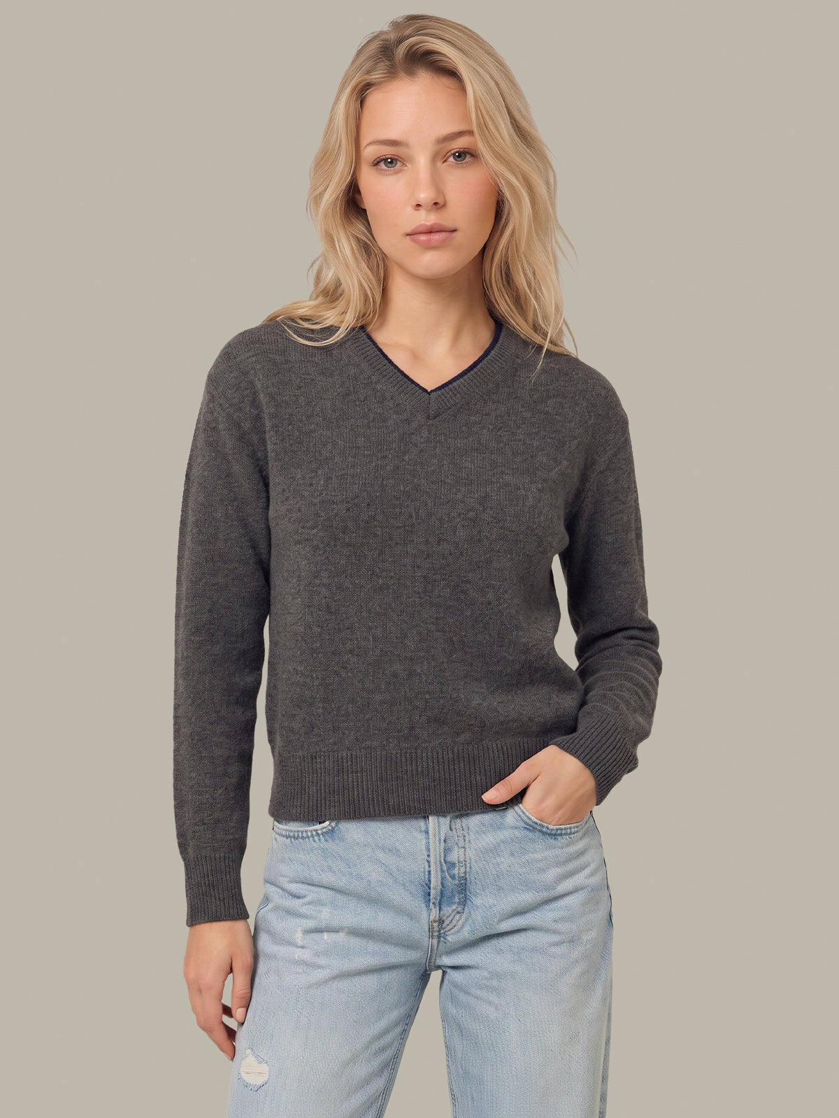 A woman modeling a dark gray v-neck knit sweater with light-wash blue jeans, standing against a solid light gray background.