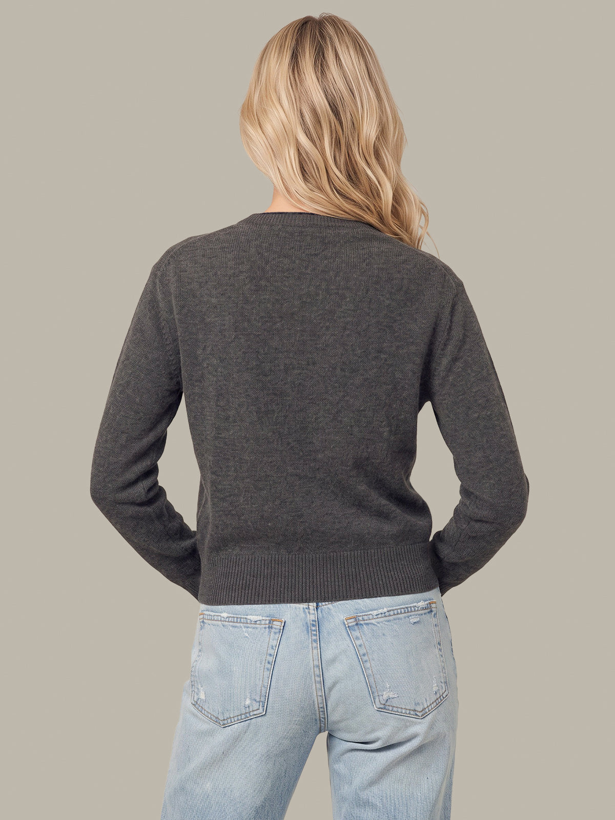A woman viewed from behind wearing a dark charcoal gray knit sweater and light blue jeans, set against a solid neutral gray background.