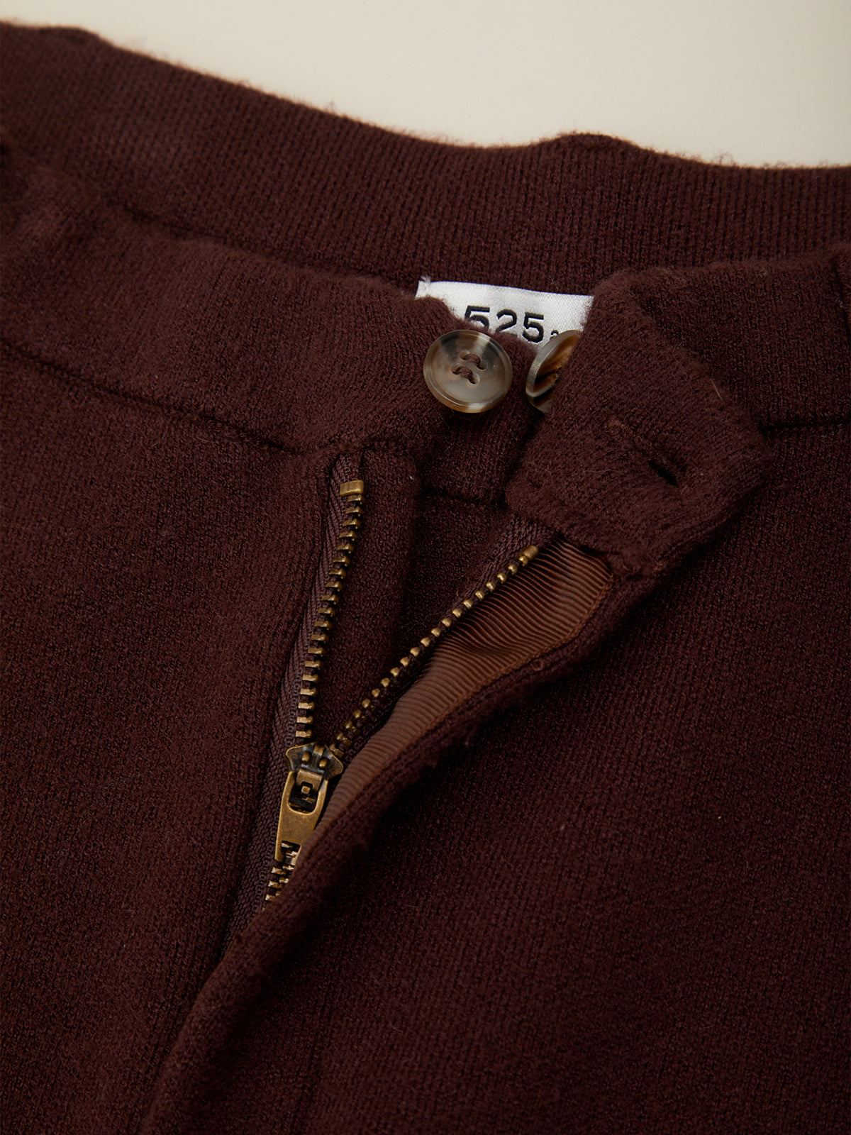 Close-up of a dark brown knit garment, showing the inner collar, a metal zipper, a button, and a small white size label.