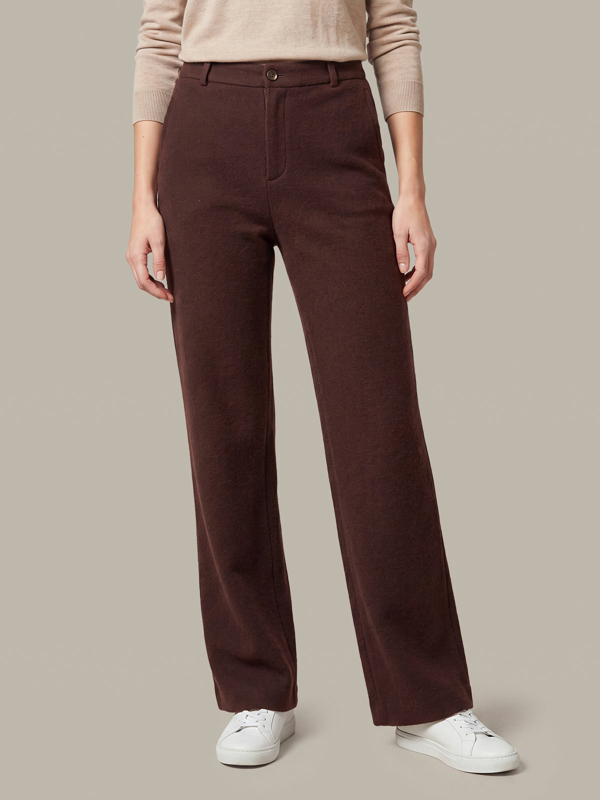 A person wearing dark brown wide-leg trousers with a beige long-sleeve top and white sneakers, shown from the waist down against a plain beige studio background.