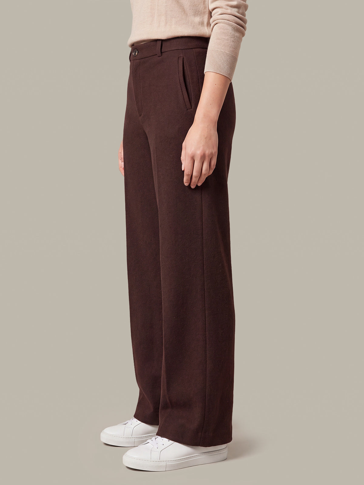 A model wearing dark brown wide-leg trousers, a beige long-sleeve top, and white sneakers, shown from the waist down against a solid beige background.
