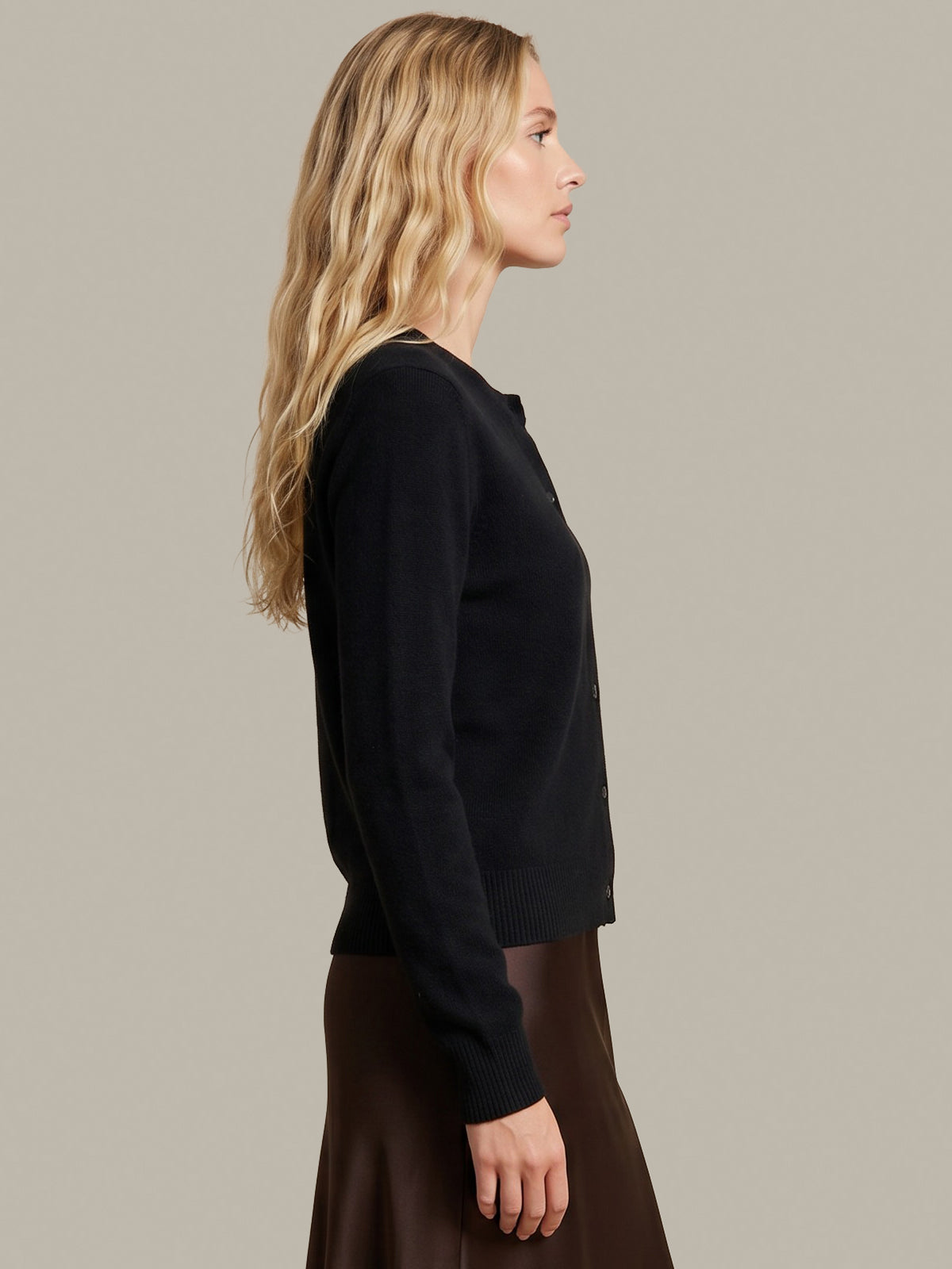 A woman in side profile wearing a black long-sleeve knit cardigan and a brown skirt, photographed against a plain warm gray studio background.