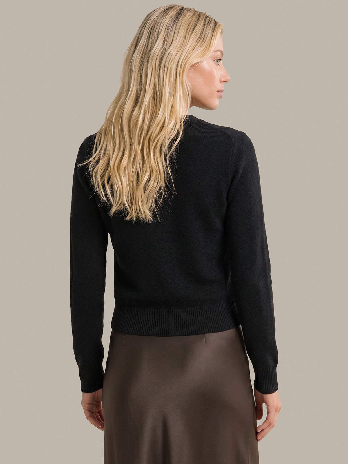 A woman with long blonde hair is shown from the back wearing a black long-sleeve knit sweater and a brown skirt, set against a solid light gray background.