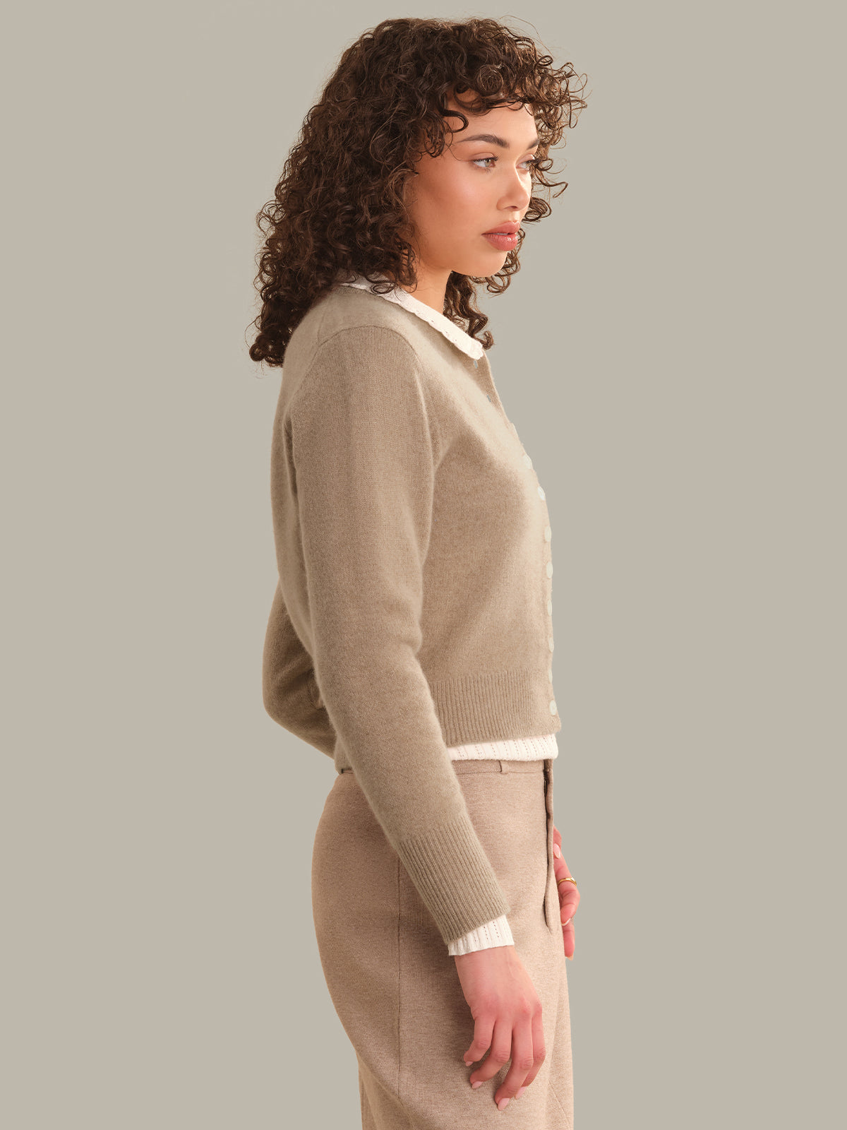 A woman in side profile wearing a beige knit cardigan with white collar and hem trim, paired with a matching beige skirt, against a solid gray studio background.