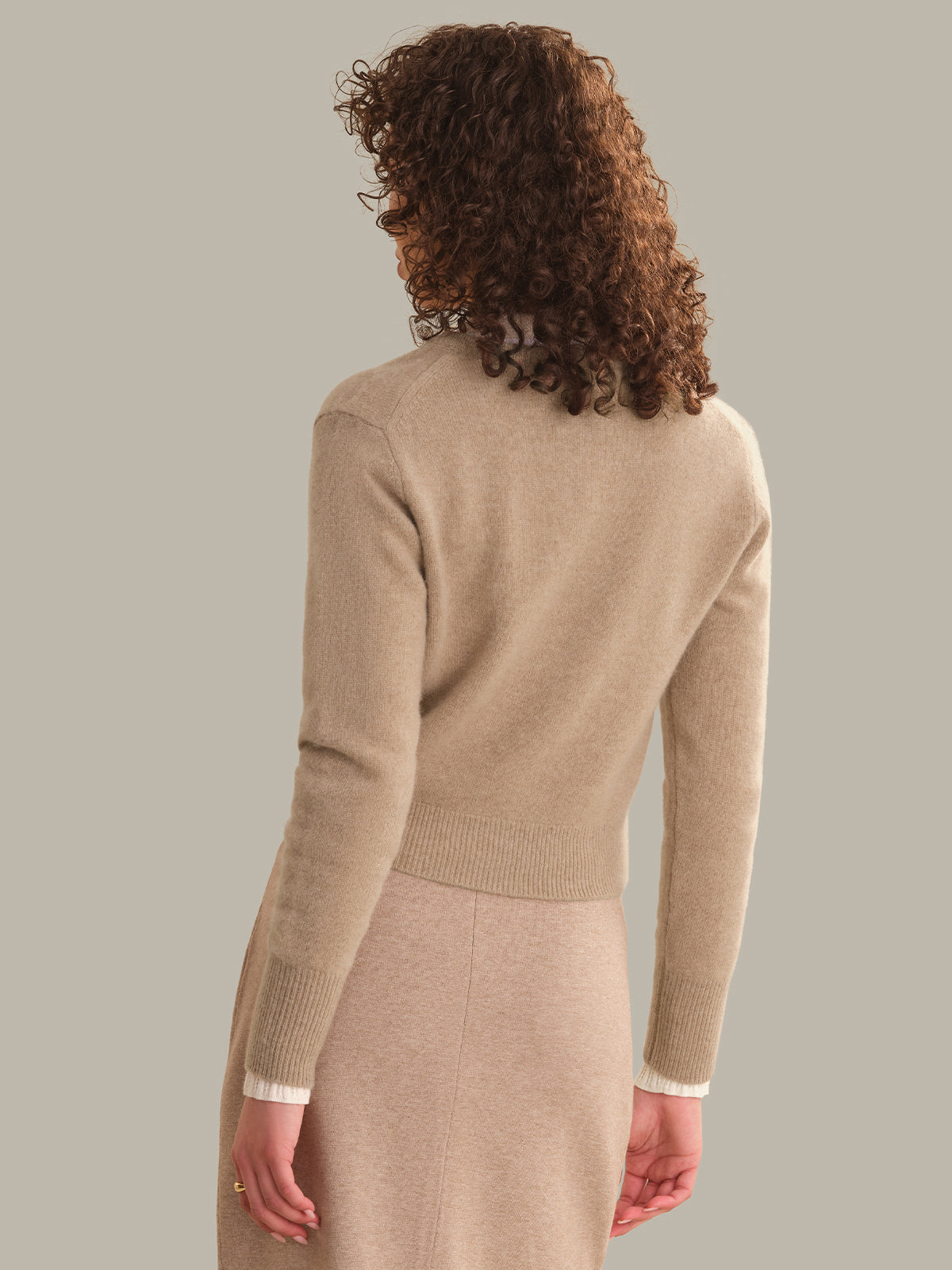 A woman with curly brown hair is shown from the back wearing a beige knit sweater and matching knit skirt against a solid taupe background.