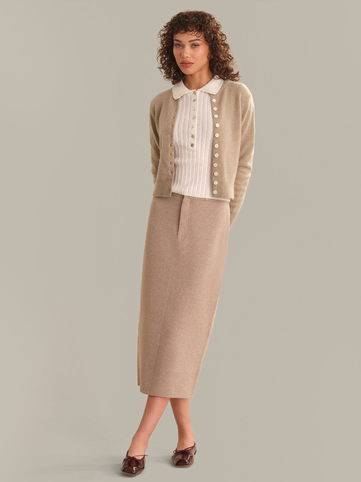 A model wearing a beige cardigan over a cream ribbed top and a matching beige midi skirt, paired with brown shoes, against a solid light gray background.