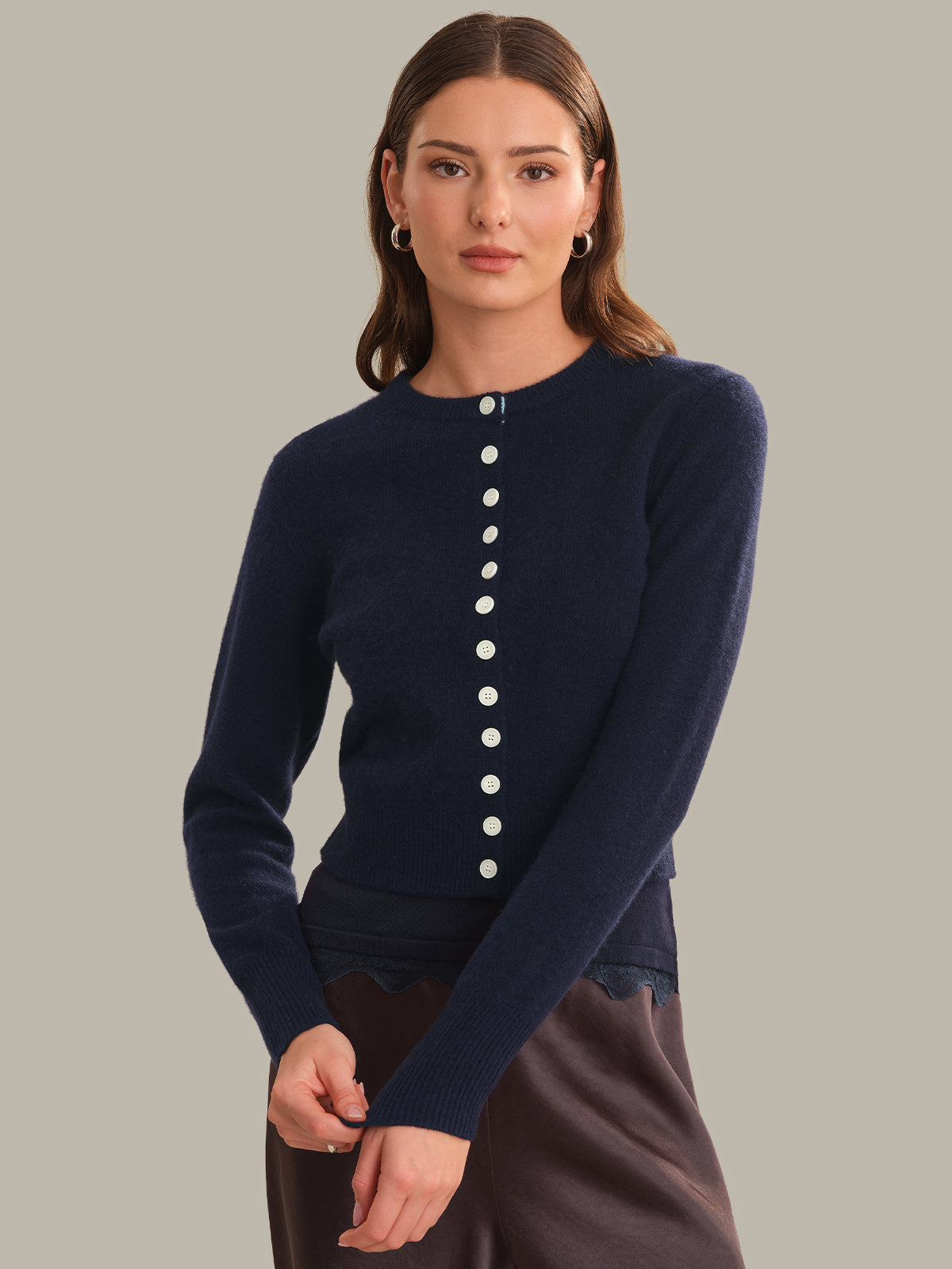 A woman modeling a navy blue long-sleeve knit sweater with a vertical row of white buttons down the front, paired with dark brown bottoms, against a solid light gray background.